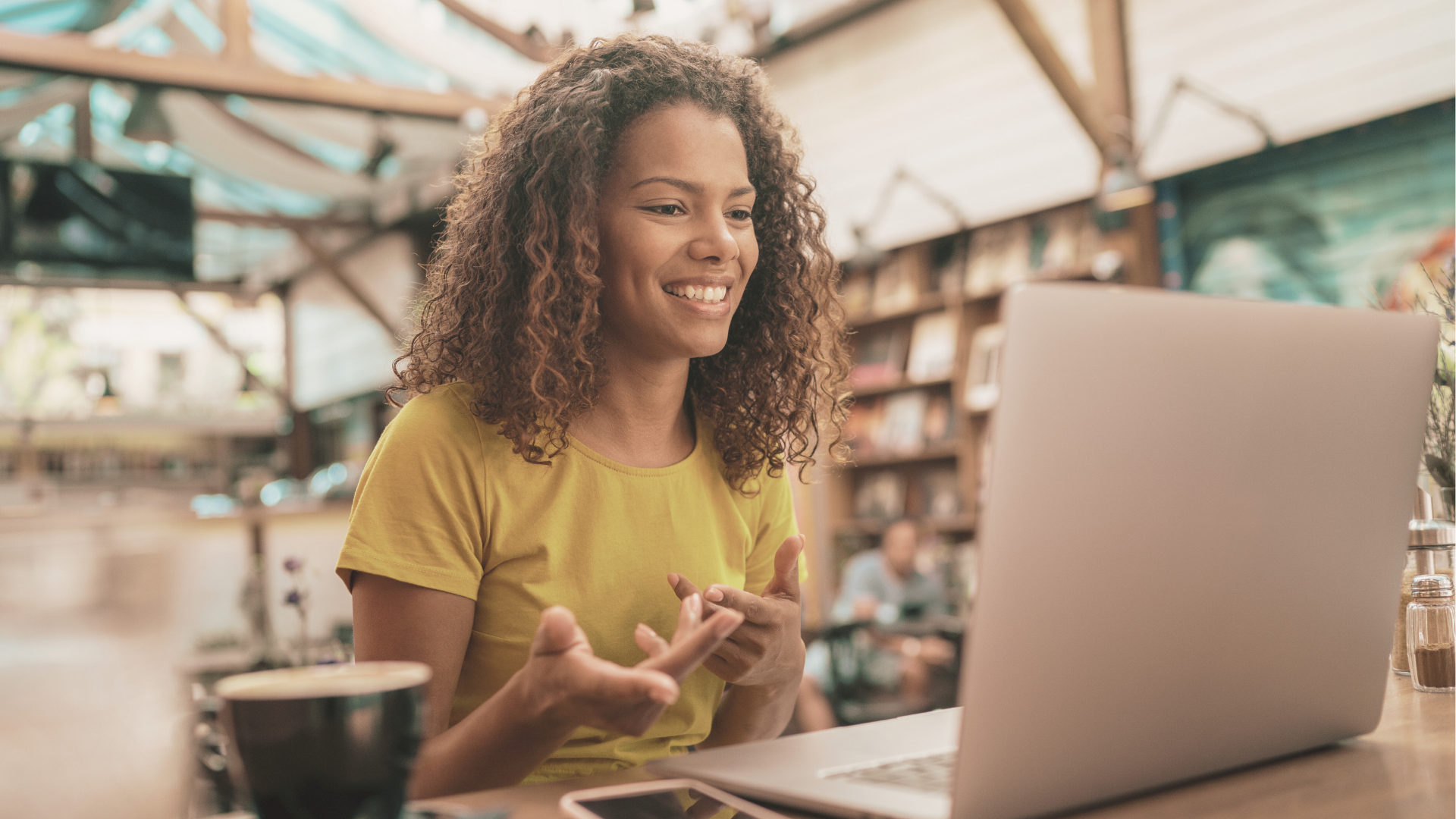 A woman with curly hair smiling at her laptop in a cafe or bookstore.