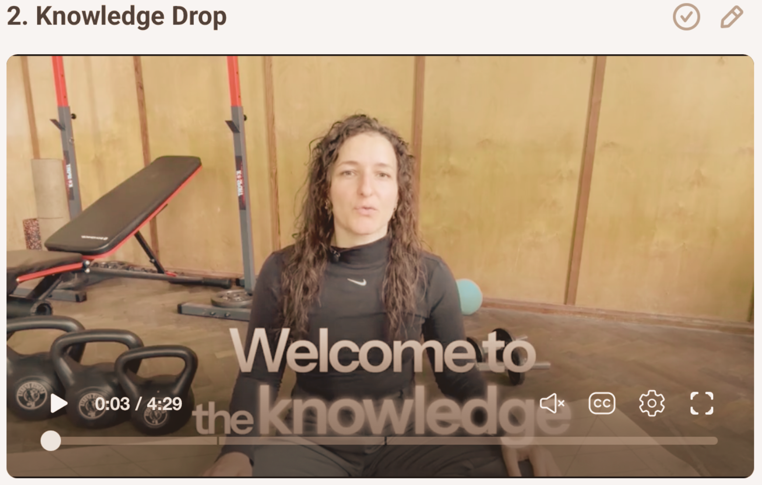A woman with curly hair sitting on the floor of a home gym space with wooden walls. Exercise equipment such as kettlebells, a weight bench, and a ball are visible. The text overlay says, 'Welcome to the knowledge'.