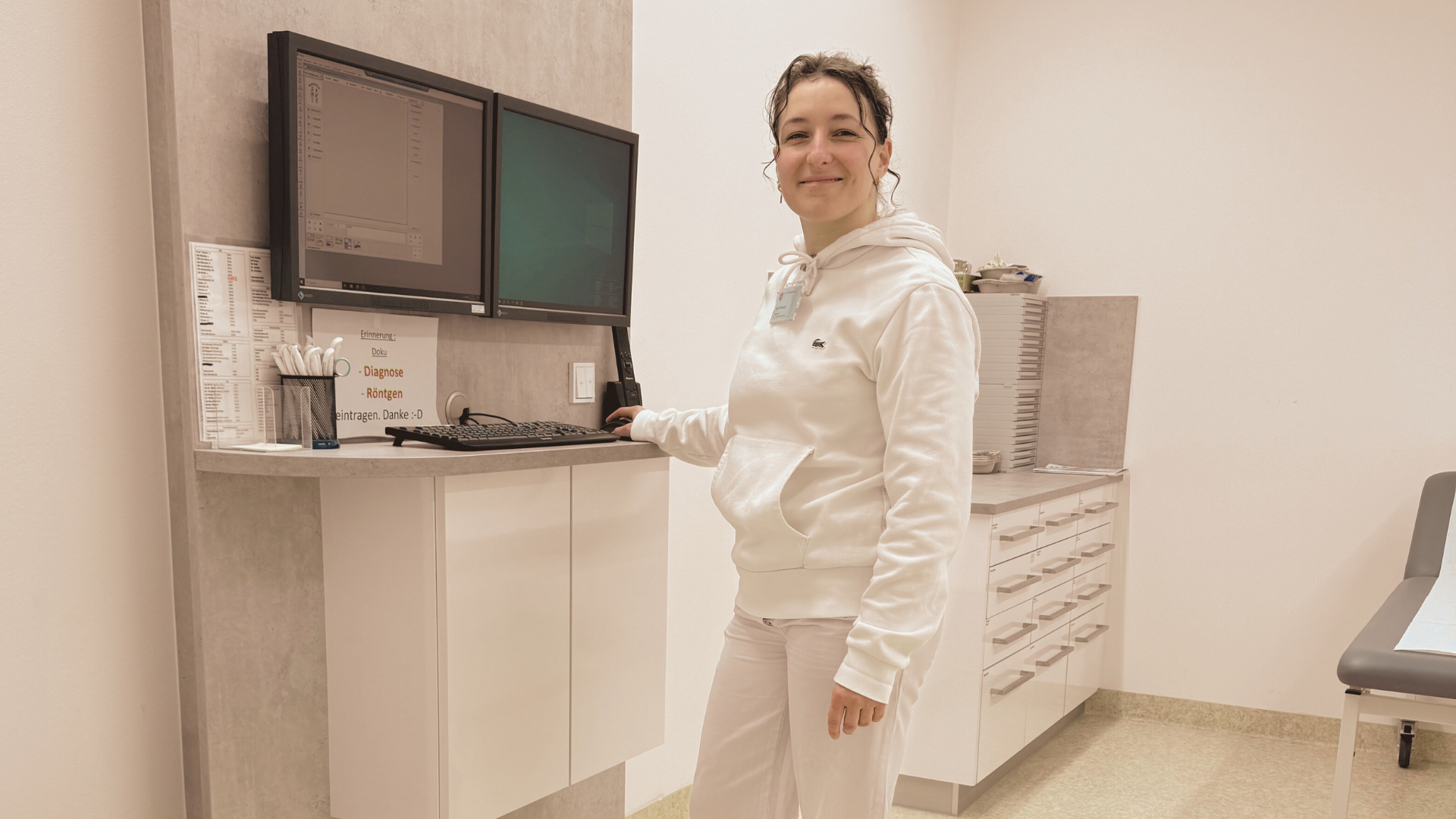 A woman in white medical attire standing in a clinical setting, using a computer with two monitors, next to a sign with German text about diagnosis and X-ray procedures.