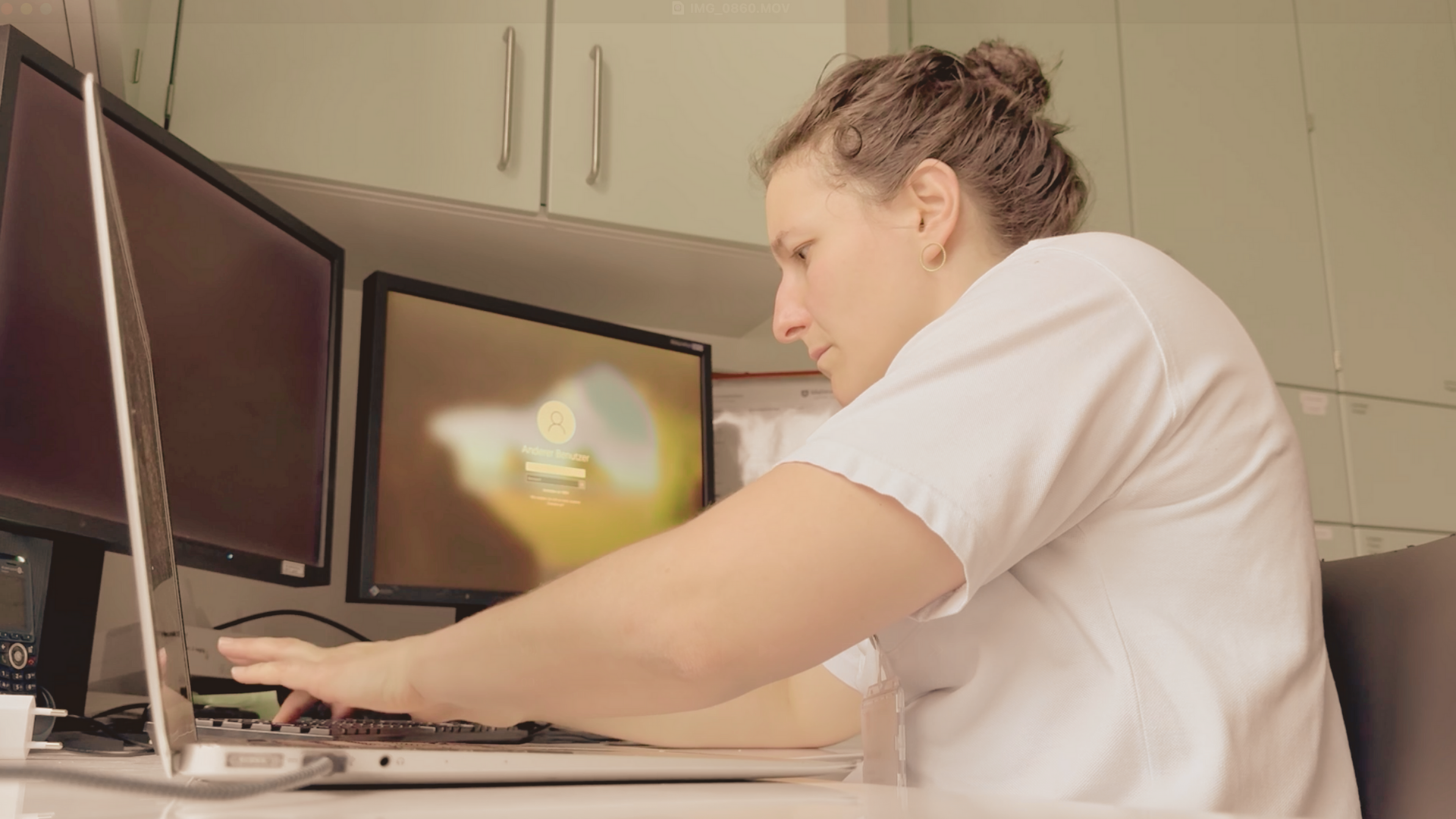 A woman with brown hair in a bun, wearing a white shirt, working with a computer in an office or workspace. She is focused on the keyboard with multiple monitors in front of her.