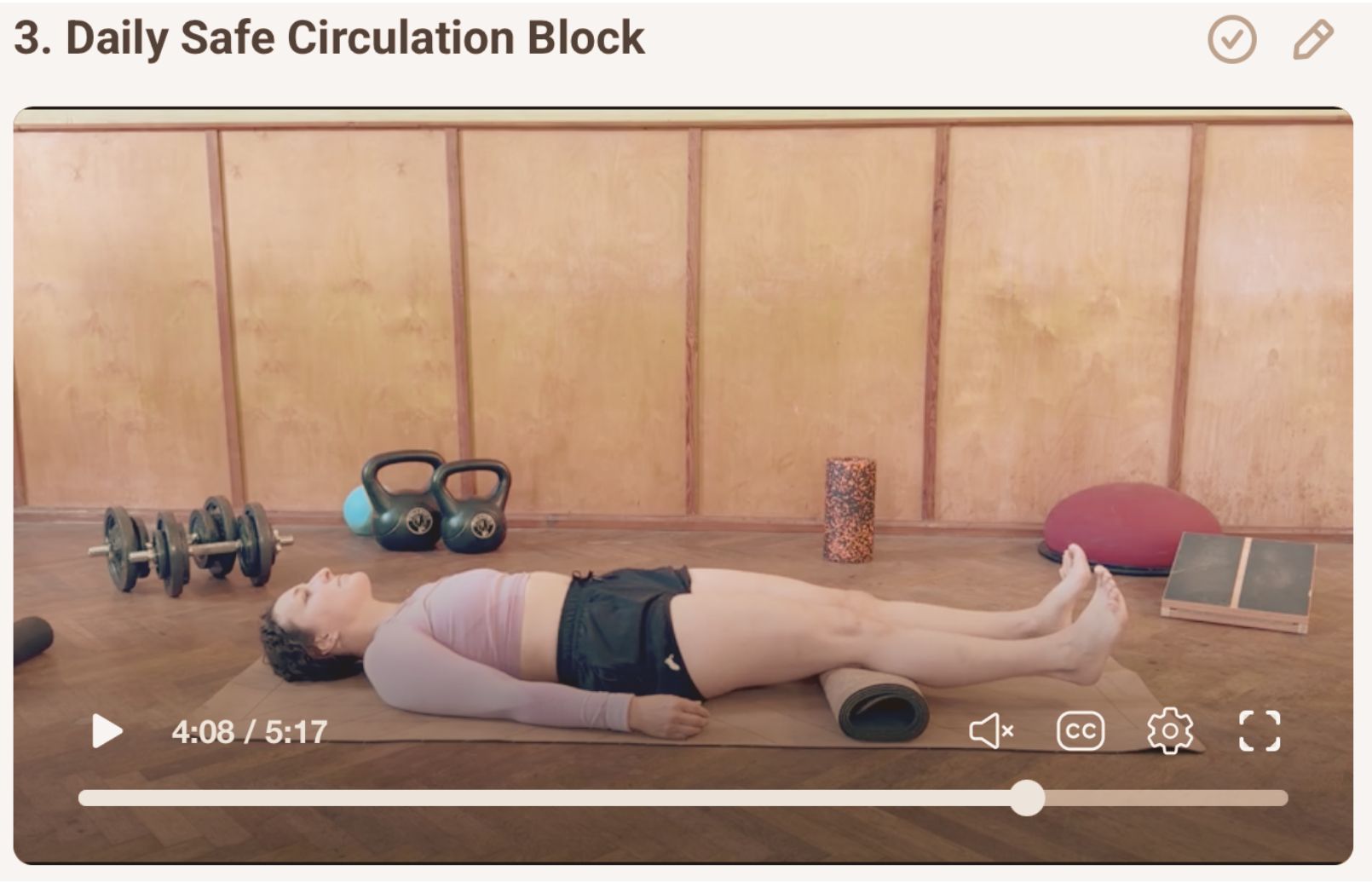 Woman lying on yoga mat in gym with kettlebells, dumbbells, foam roller, balance disc, and exercise blocks in the background.