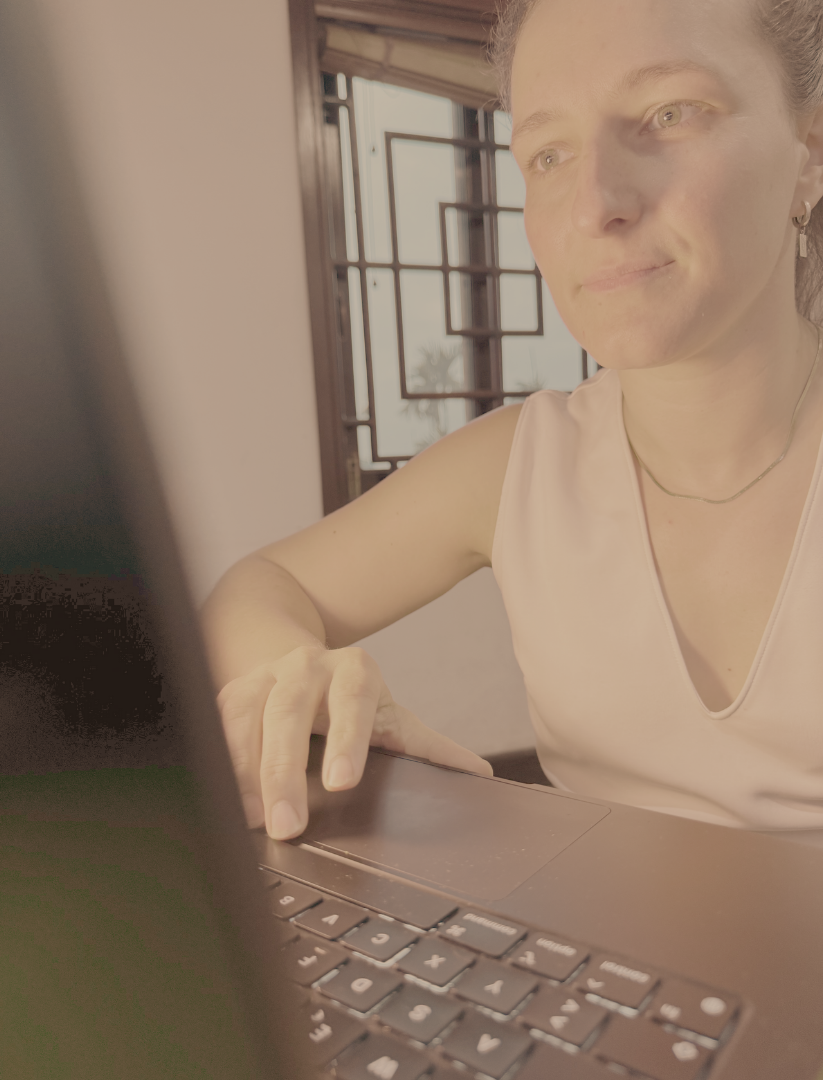 A woman with light skin sitting at a desk, using a laptop, with sunlight coming through a window behind her.