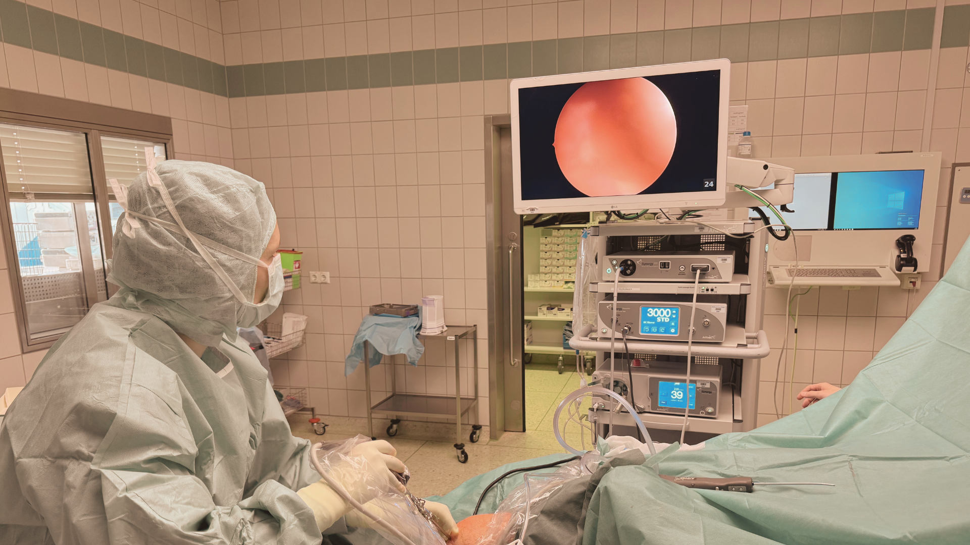Medical professional in surgical gear performing a procedure in an operating room with monitors and medical equipment.