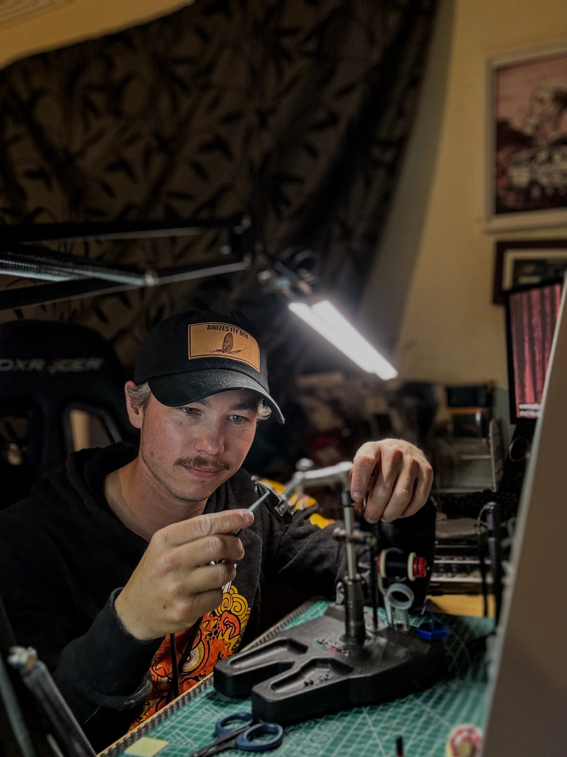 A man with a mustache and wearing a black cap and hoodie working on a model or small mechanical project at a cluttered workbench, with various tools and equipment around him.