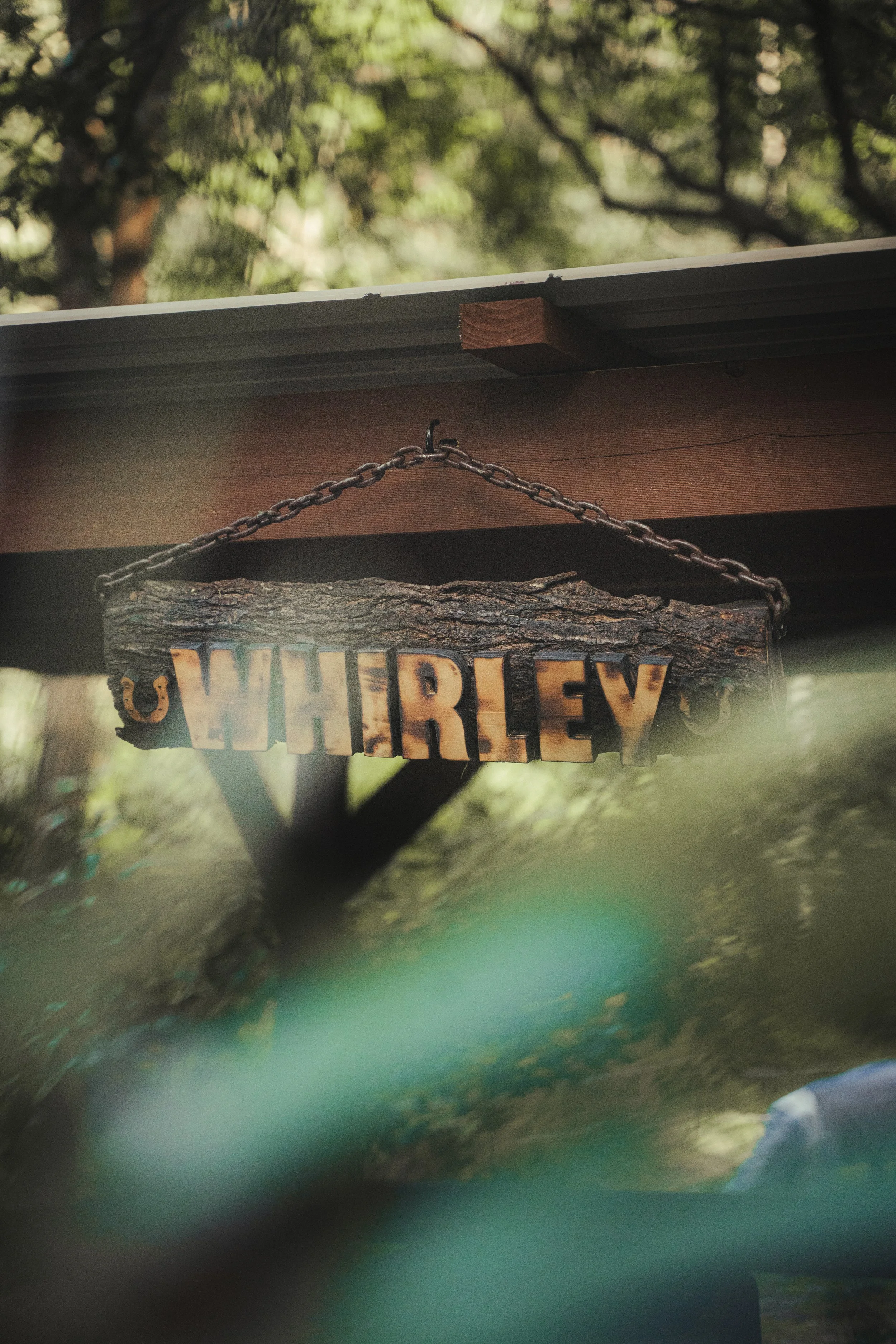 A wooden hanging sign spelling "WHIRLEY" attached to a chain, mounted under the edge of a wooden overhang, with trees and greenery in the background.
