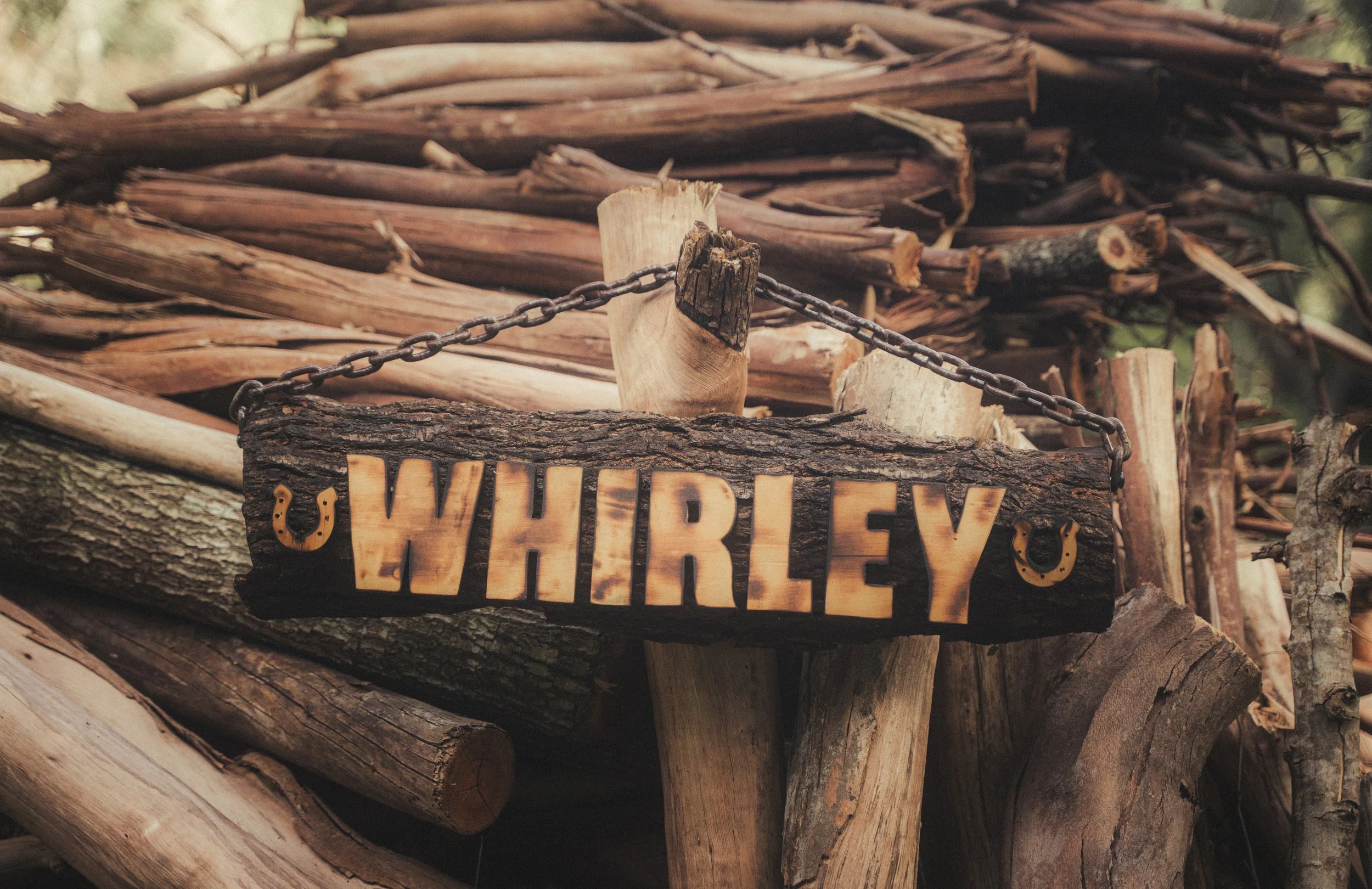 A weathered wooden sign with the name 'WHIRLEY' hangs from a chain in front of a large pile of chopped firewood.
