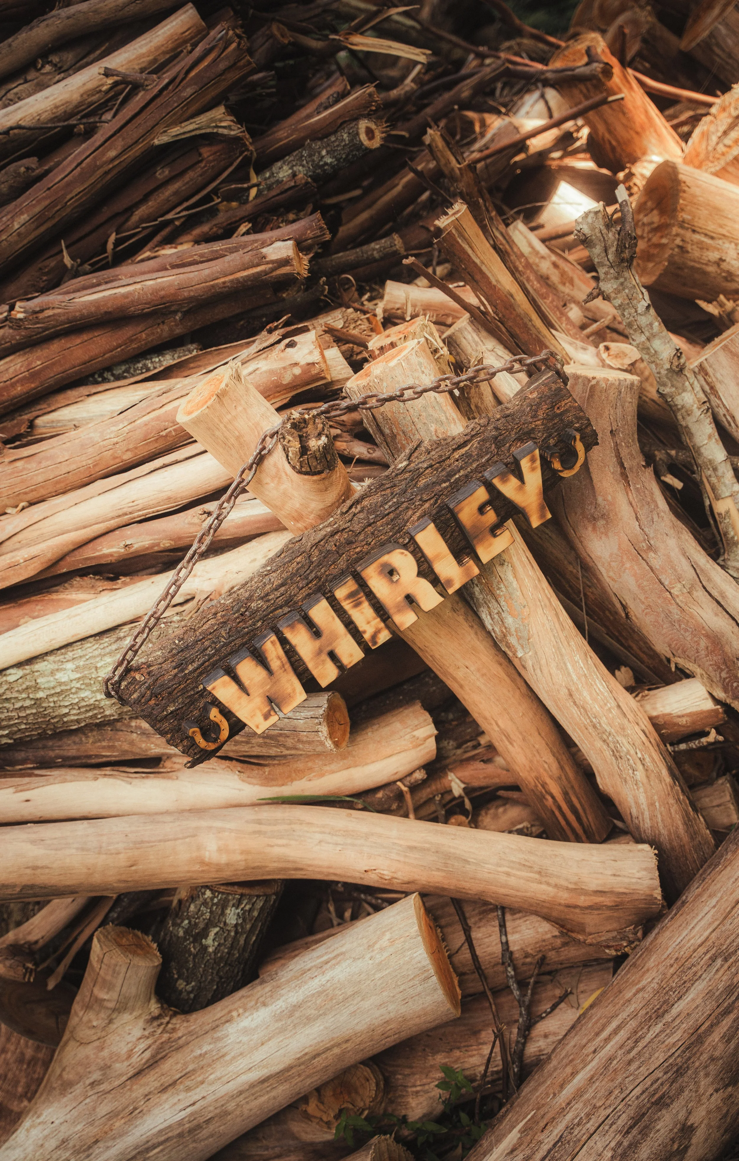 Stack of chopped firewood with a wooden sign labeled 'WHEELER'S' hanging on a chain.