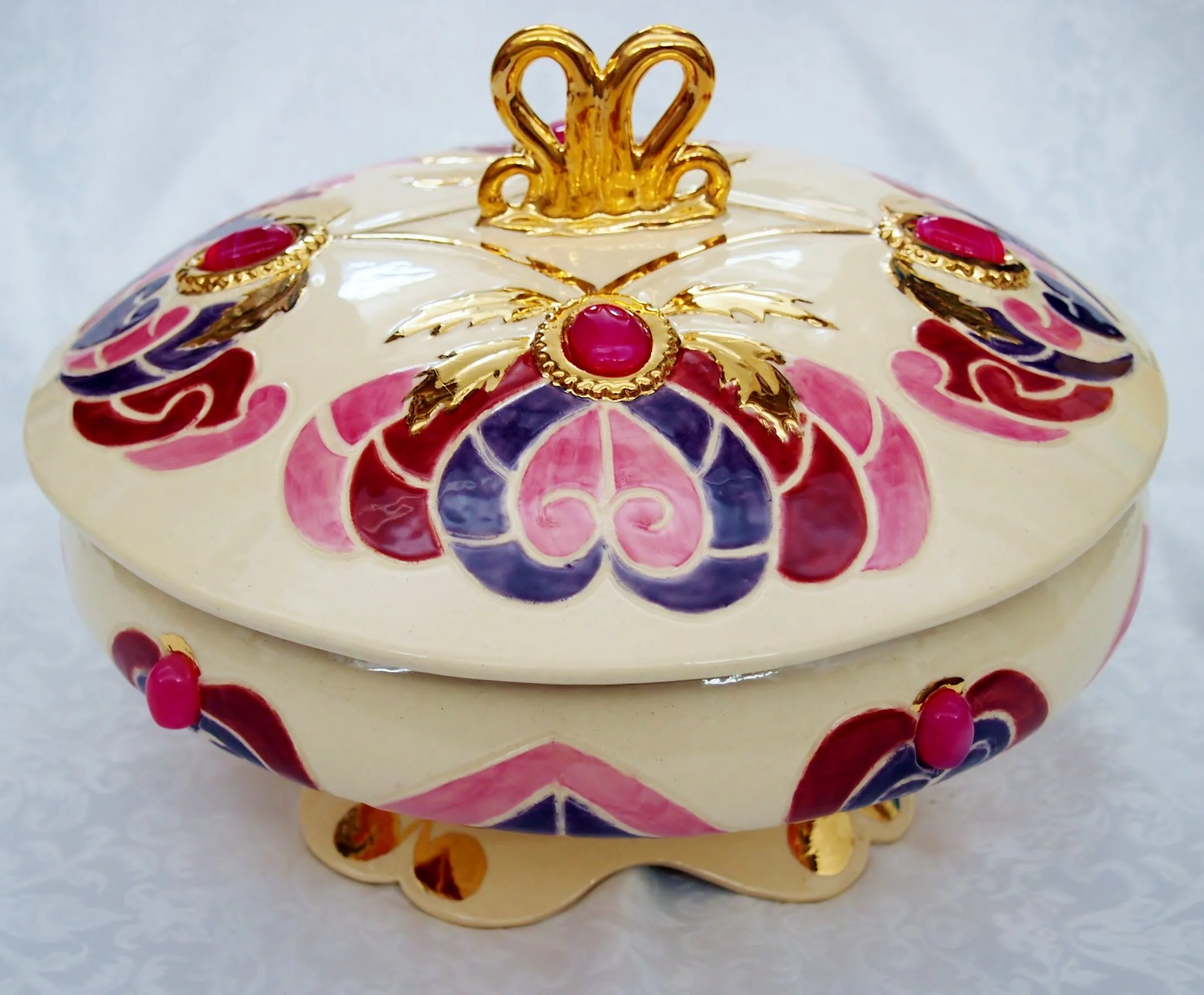 A decorative ceramic box with a lid, embellished with pink, purple, and gold patterns, featuring a gold crown-like ornament on top, resting on a white surface.