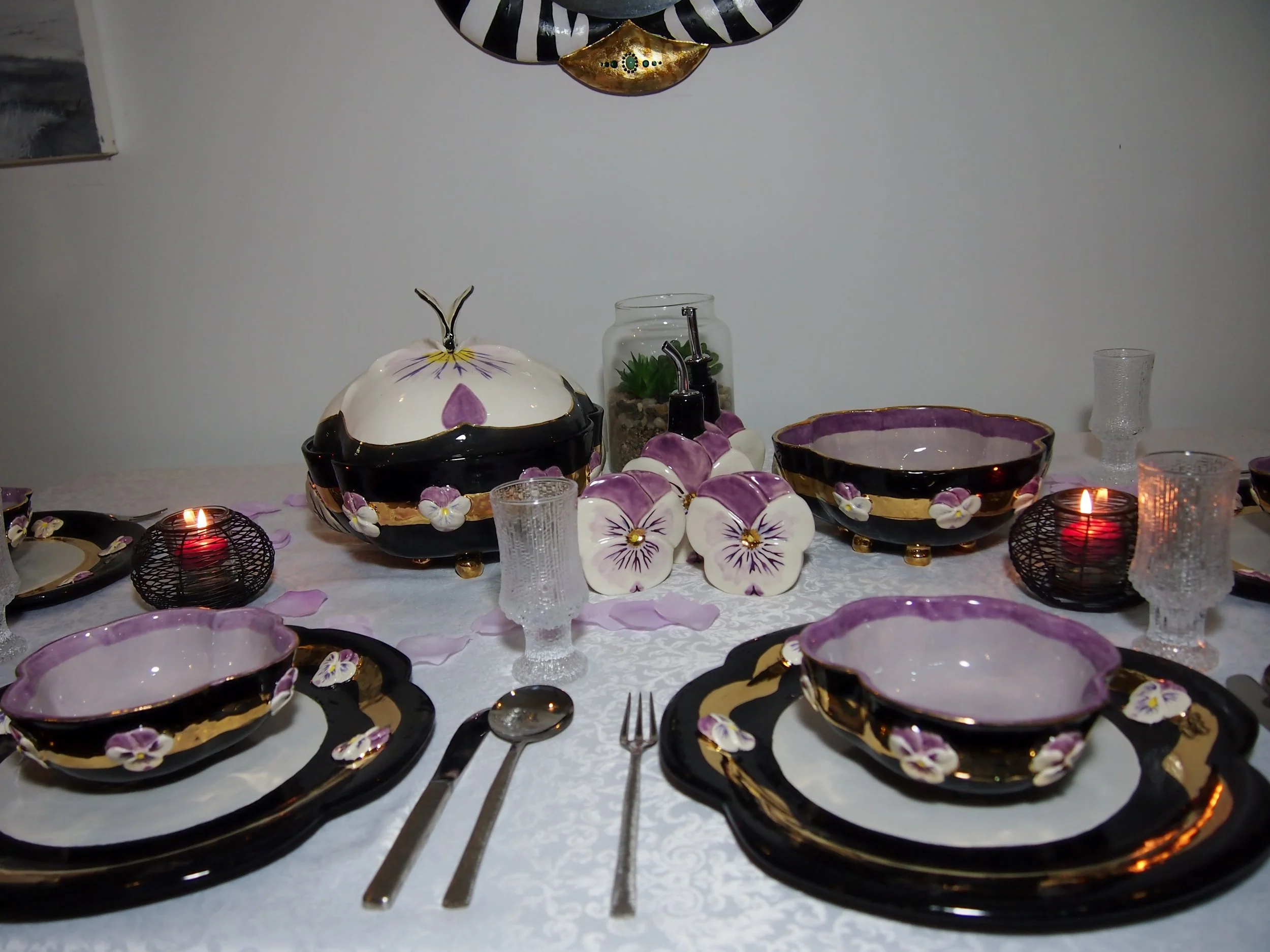 Decorative dinnerware set with black, gold, and purple floral designs, including bowls, plates, and matching lids, arranged on a white tablecloth with candles and glassware.