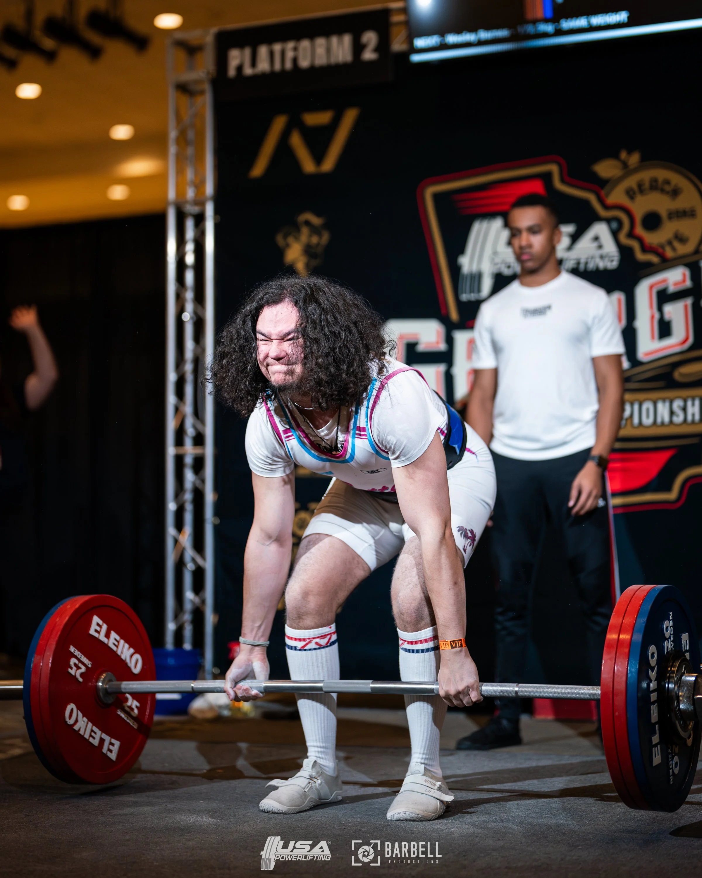 A man with long curly hair and a scrunched face, wearing a white athletic outfit with colorful trim, performing a deadlift at a powerlifting competition. A spotter stands behind him, dressed in black and white, watching intently. The background features a stage with a black backdrop displaying logos and text related to the event.