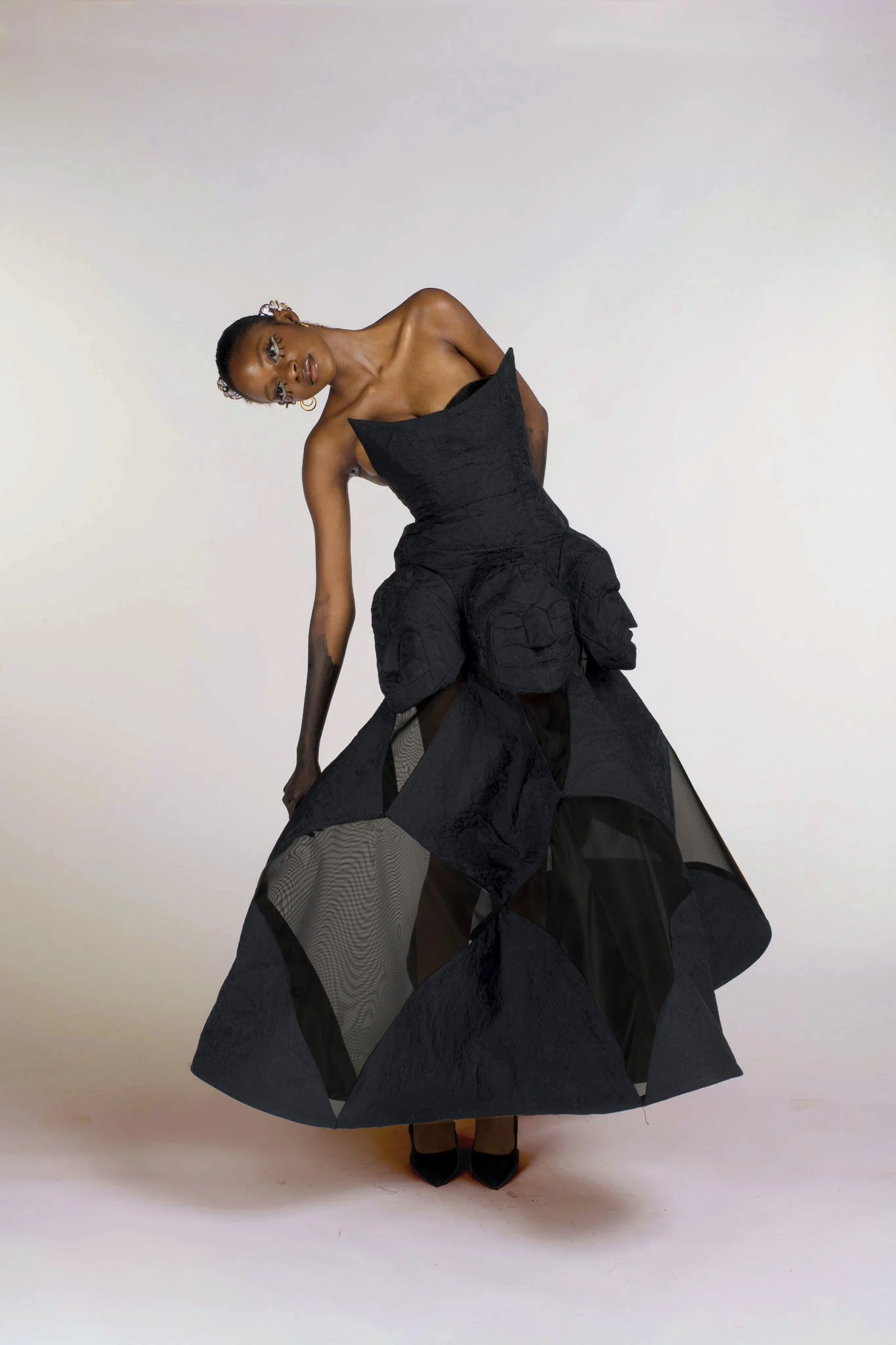 A woman wearing a black strapless dress with layered skirt and high heels, standing against a plain light background, looking at the camera with her head tilted to the side.