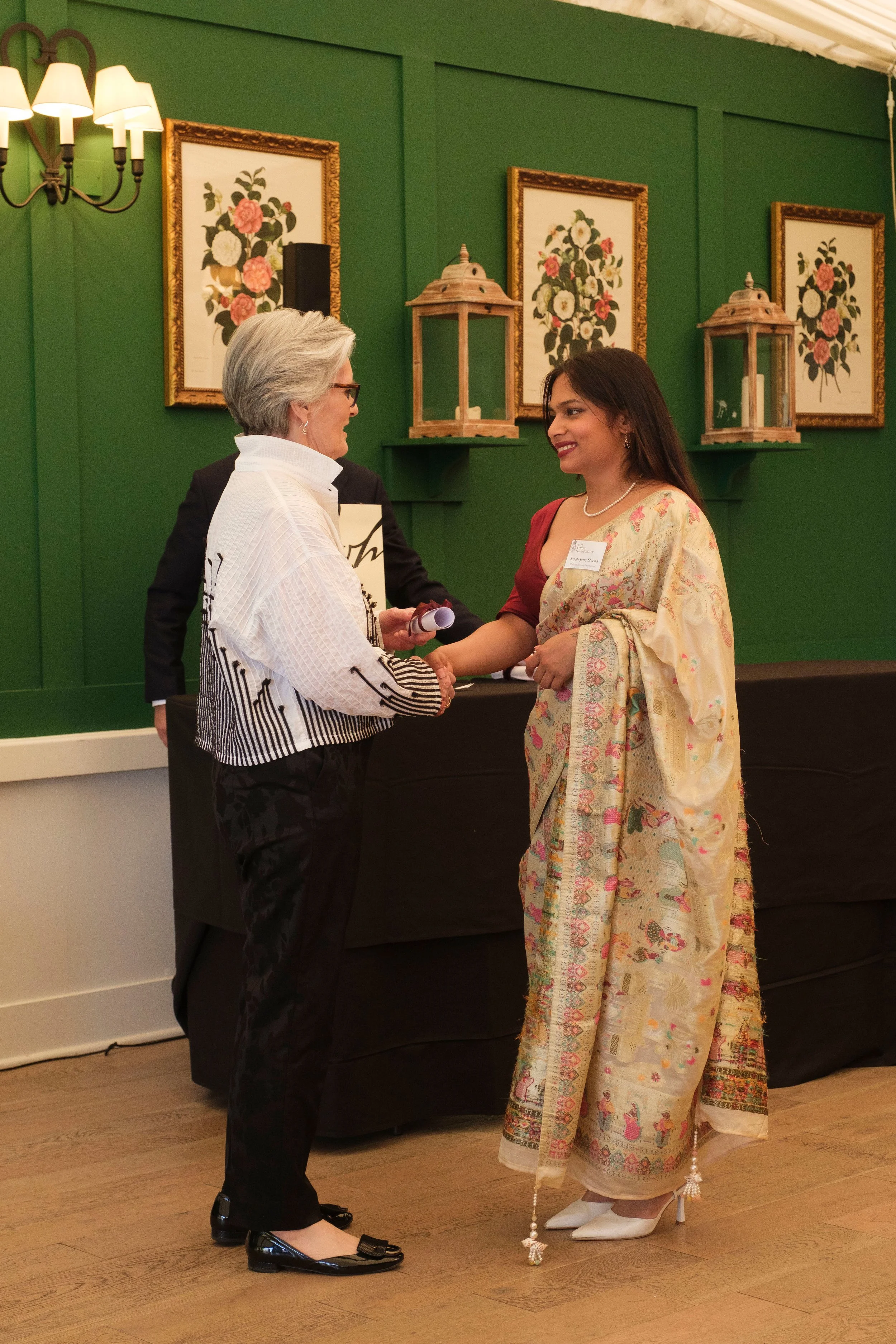 Two women shaking hands at a formal event, with one woman wearing a traditional saree and the other in a modern outfit, in front of a green wall with framed floral artwork.