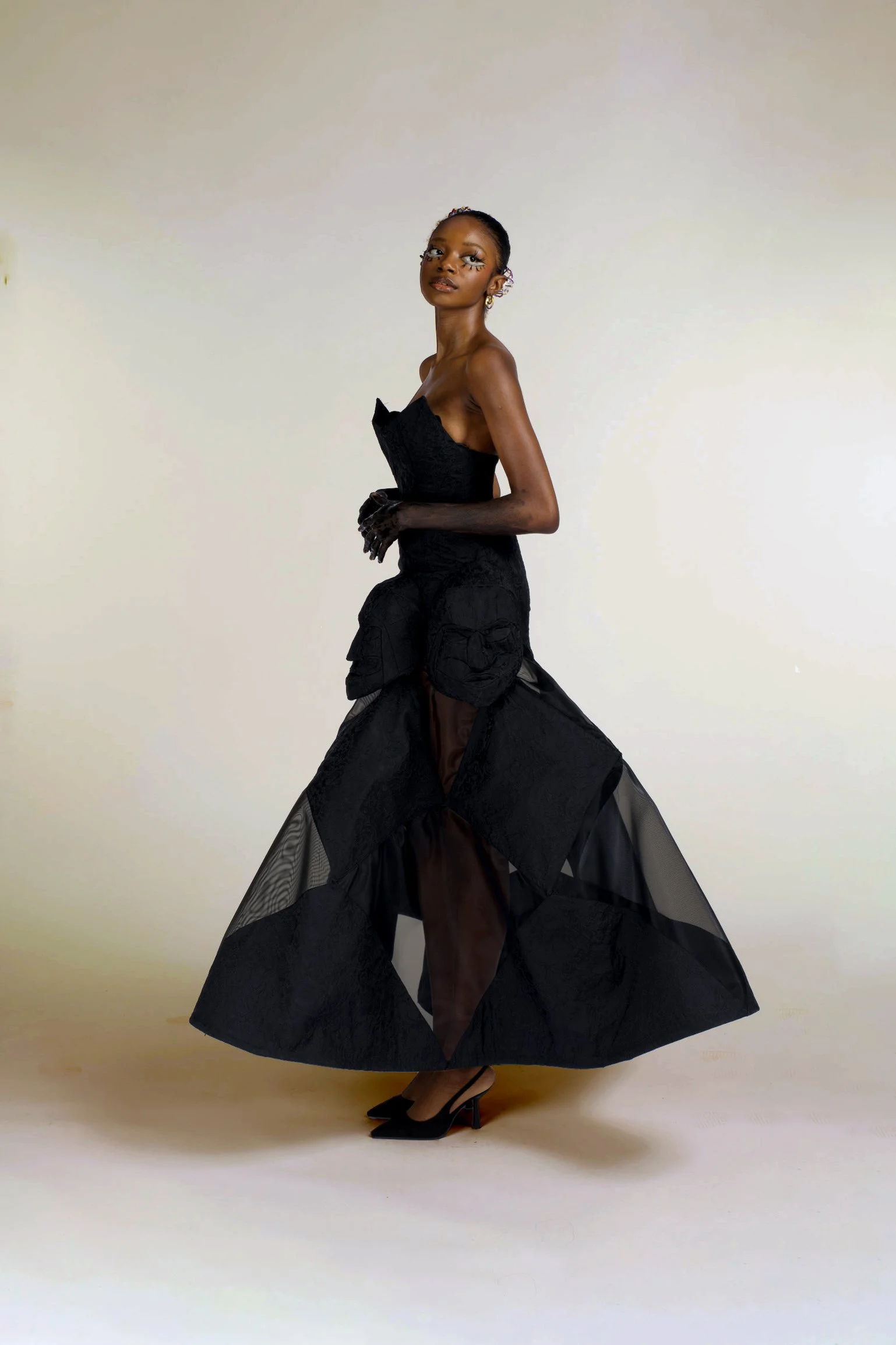 A woman wearing a black dress with a sheer, voluminous skirt and high heels, standing against a plain light-colored background.