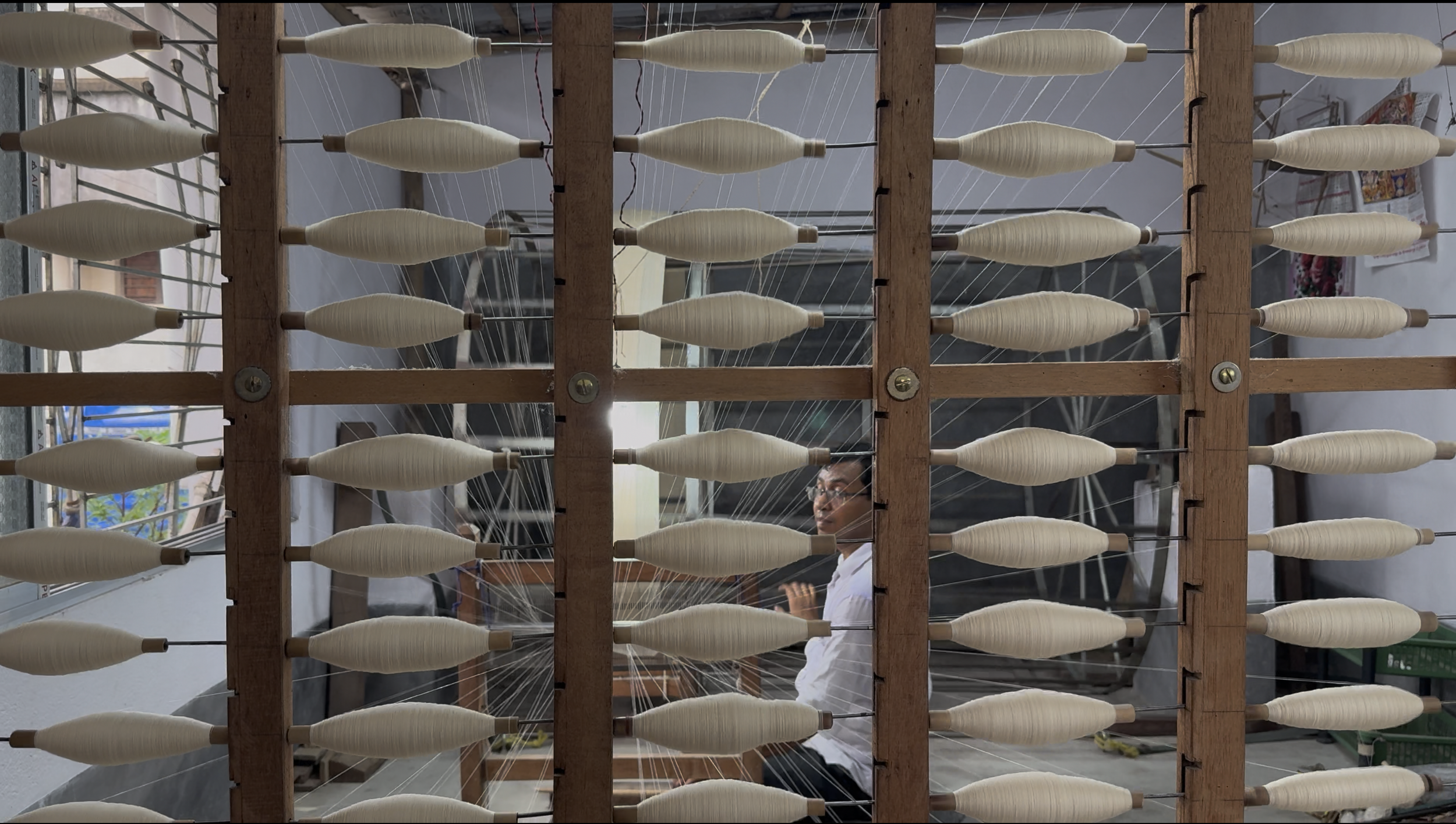 A person sitting behind a large wooden loom, weaving white thread into fabric, with multiple spools of thread arranged on the loom.