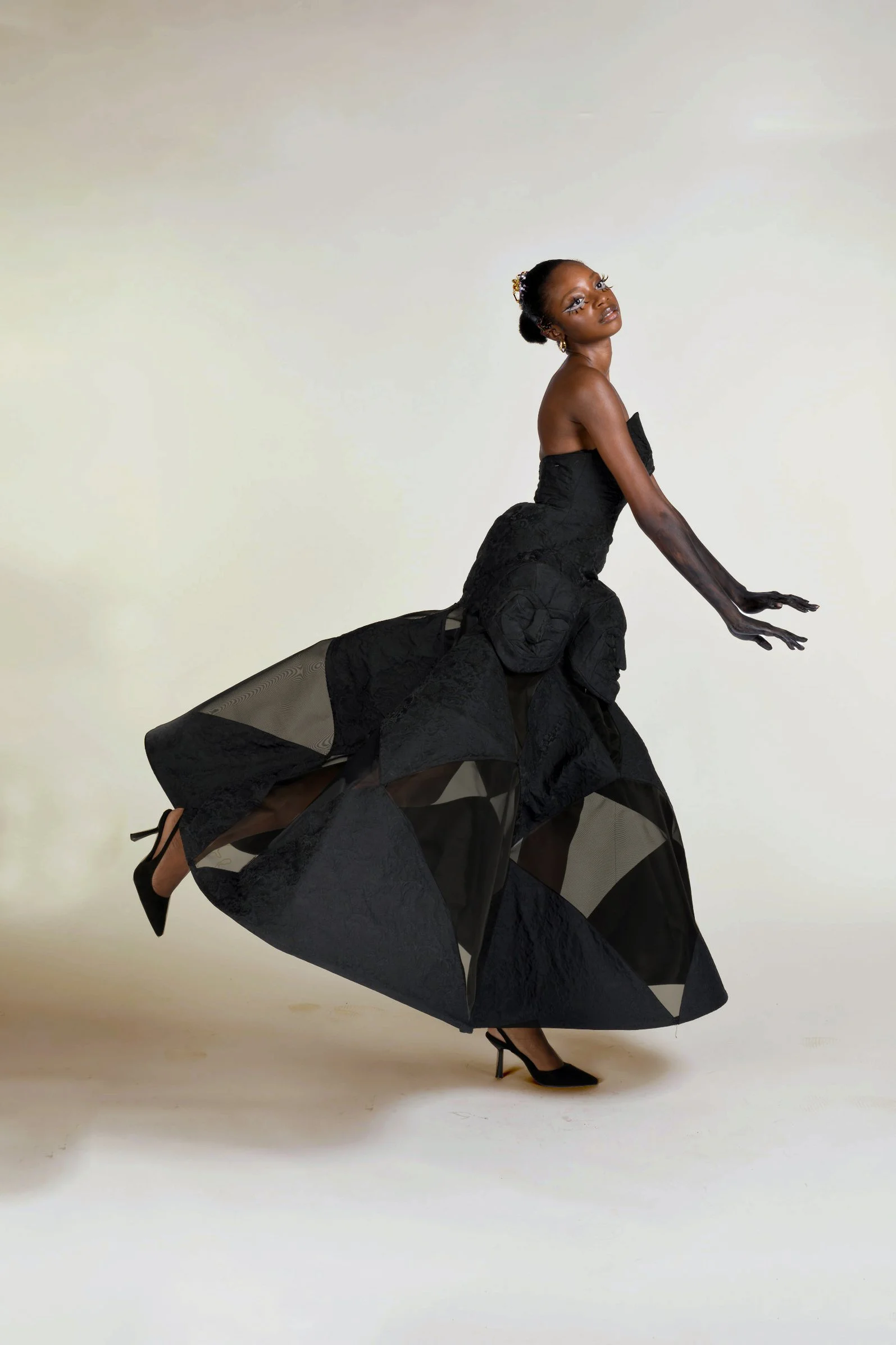 A woman in a black, intricate dress with sheer and opaque sections poses gracefully against a plain backdrop, wearing black high heels and accessorized with earrings and hair ornaments.