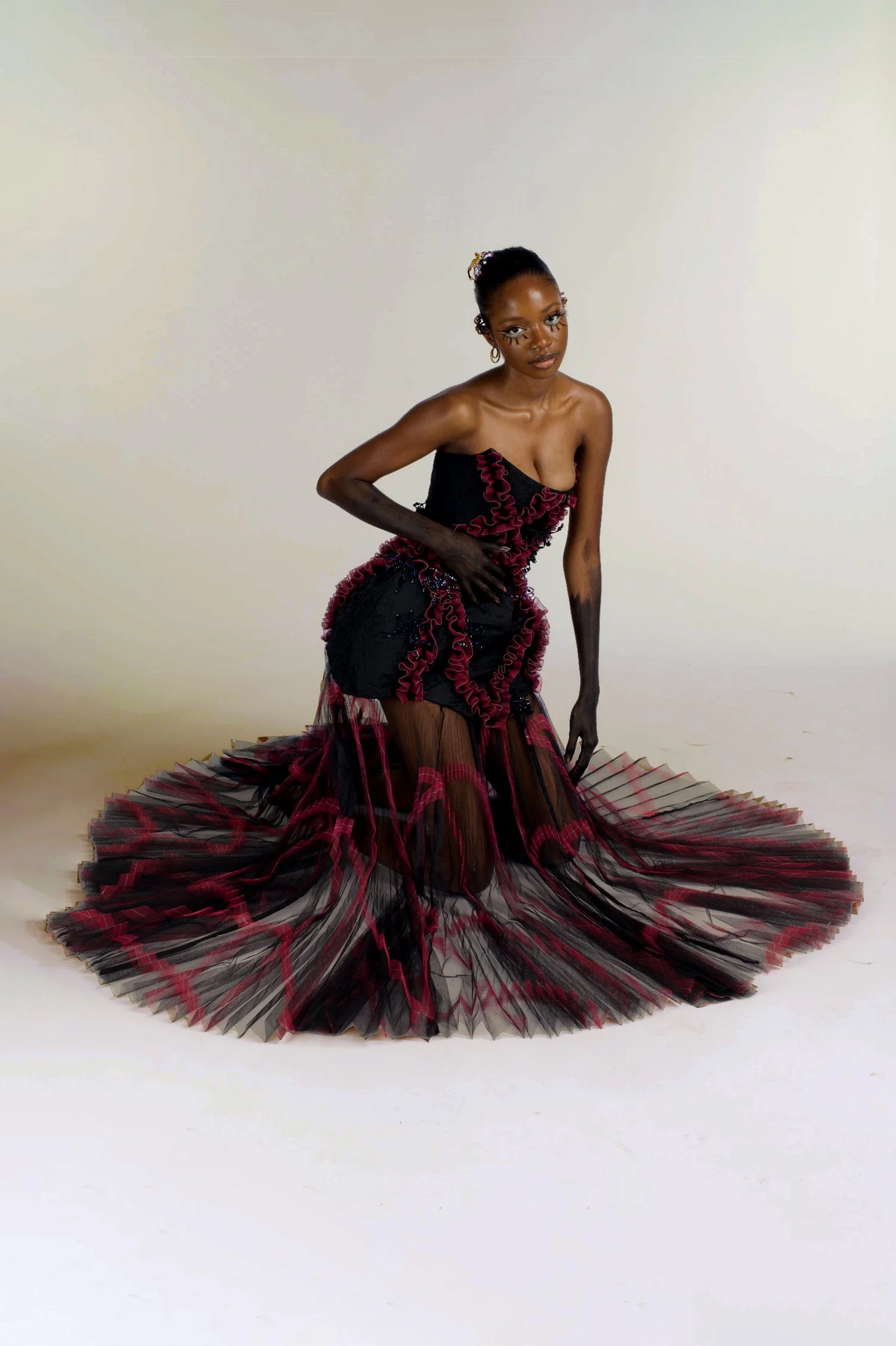 A woman wearing an elegant black and red ruffled gown with a long, pleated, sheer skirt sitting on a plain studio background.