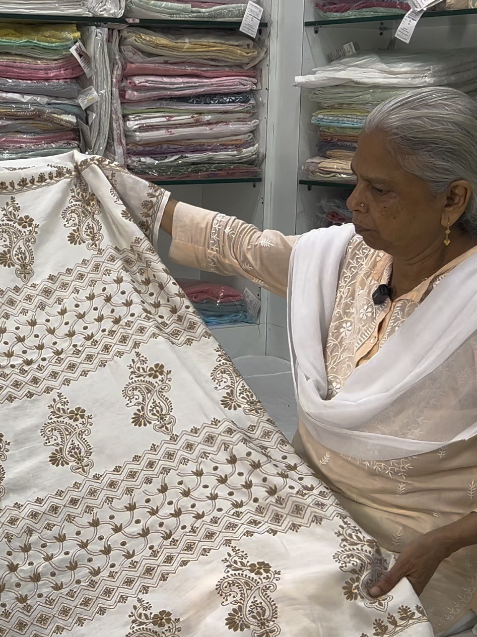 An elderly woman in traditional Indian dress inspecting a white embroidered fabric with gold threadwork in a fabric store surrounded by shelves of folded fabrics.