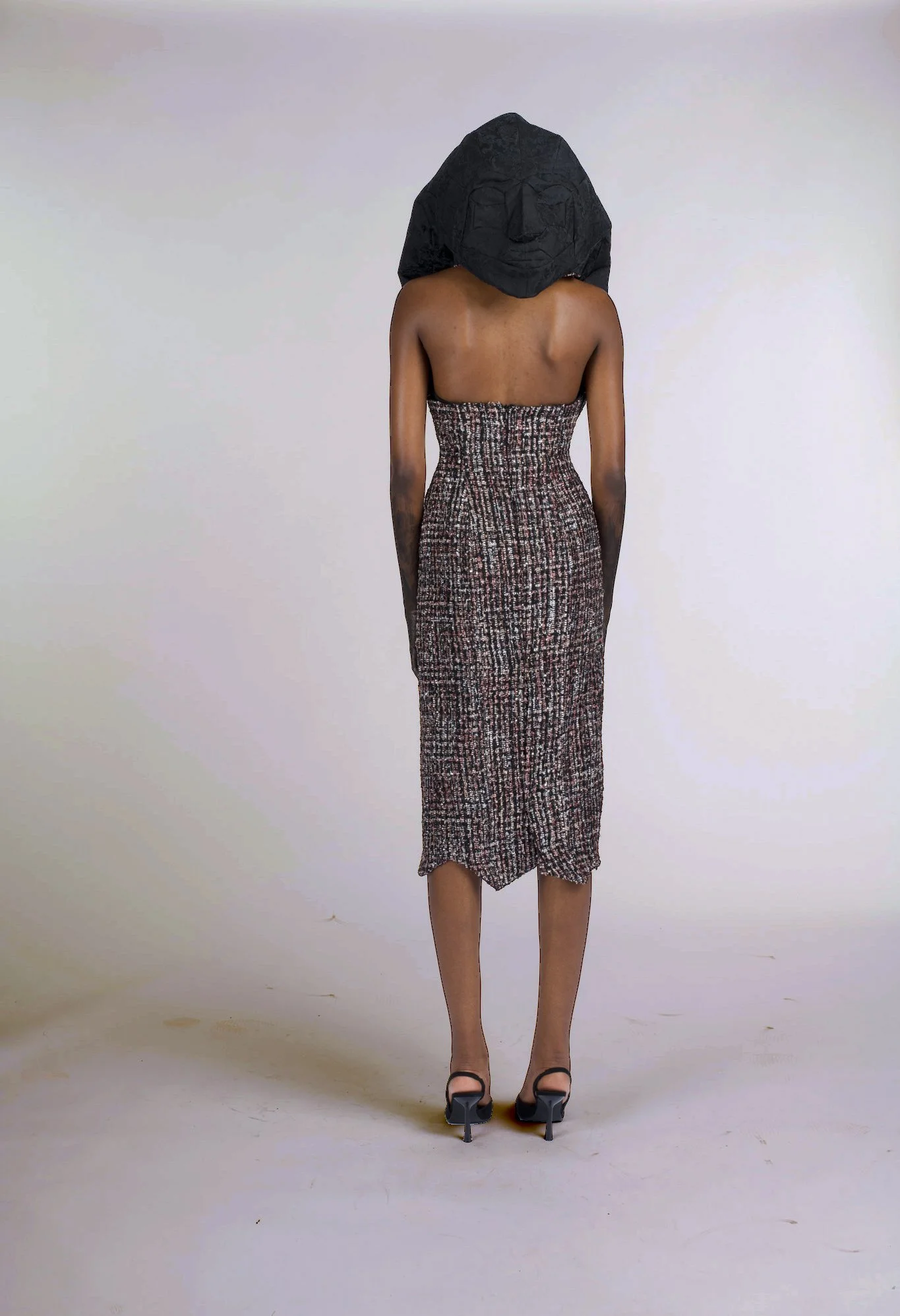 A person wearing a strapless dress with a patterned skirt and black high heels, standing face away from the camera, with a paper mask featuring a smiling face placed on their head.