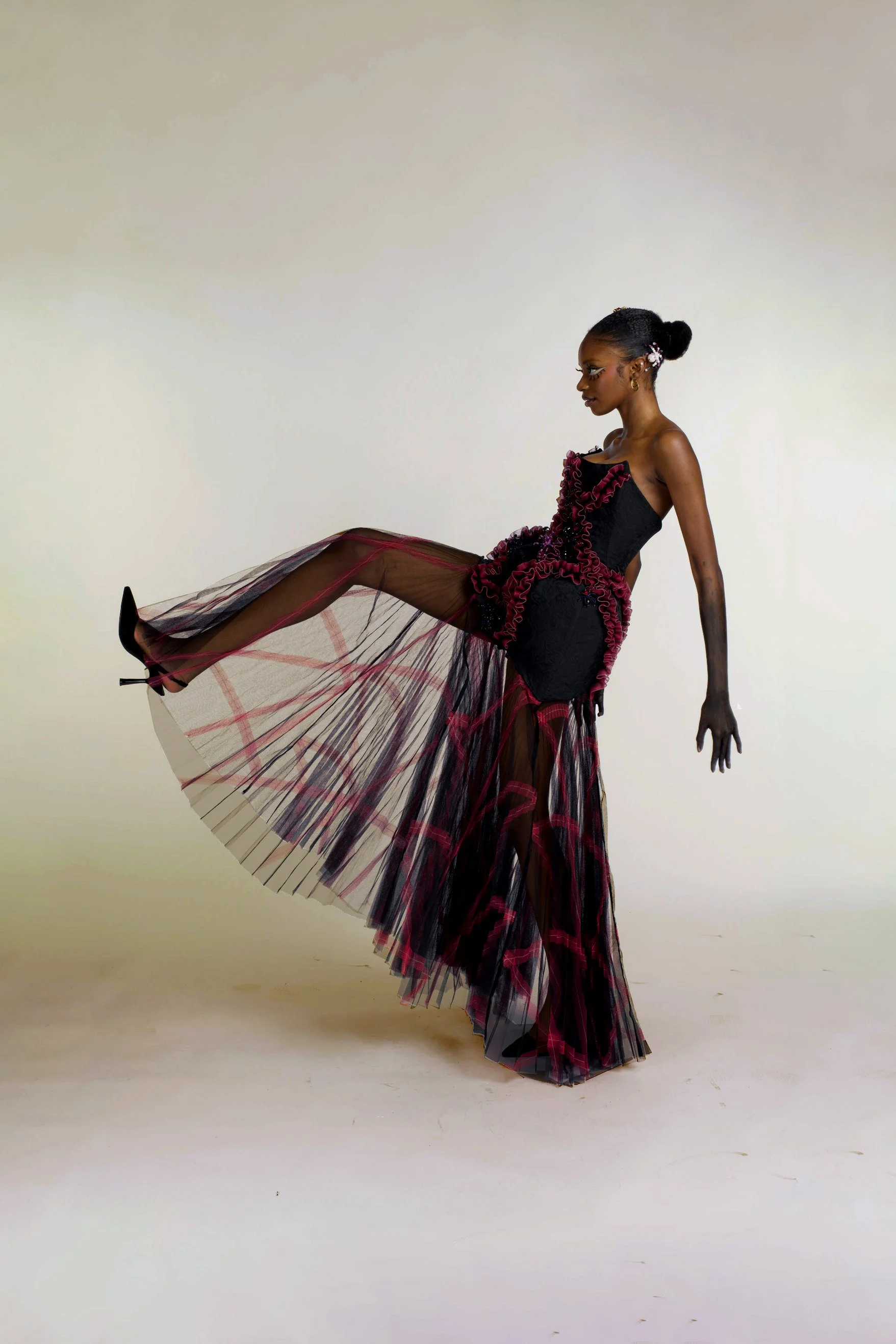 A woman in an elaborate black and red dress with a sheer, pleated skirt lifts her leg forward in a pose against a plain light background.