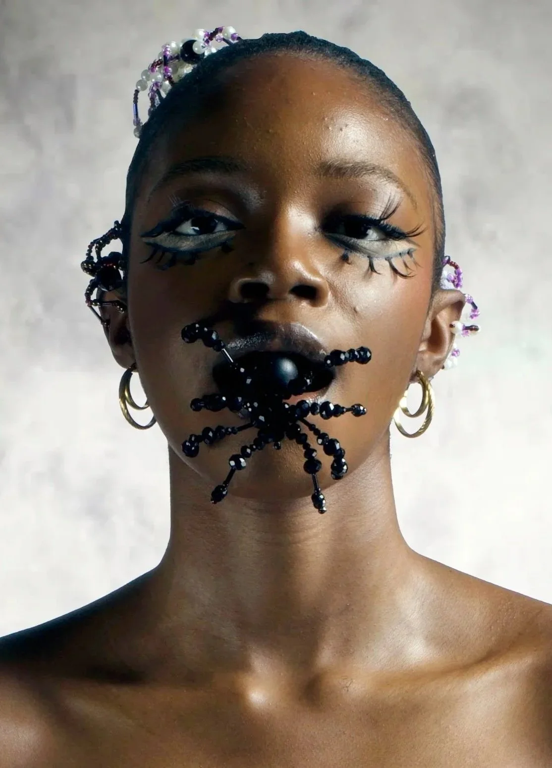 A woman with artistic makeup, including drawn eye and eyelash designs, and makeup on lips and chin resembling a black spider with extended legs, wearing multiple earrings and beaded hair accessories.