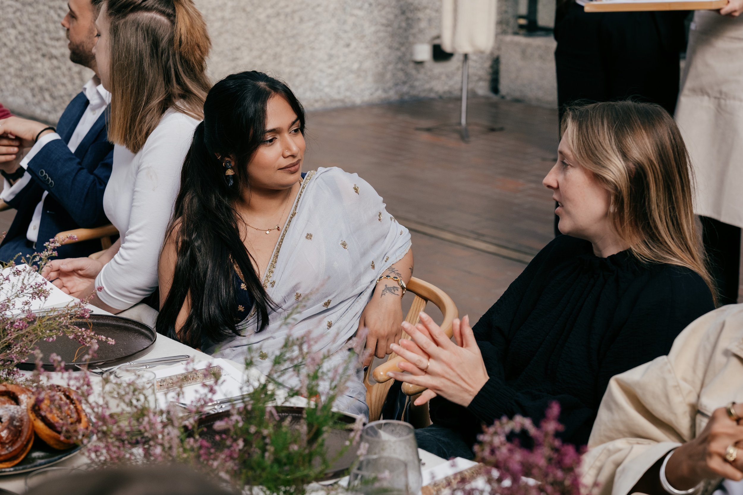 Two women engaging in conversation at a dining table decorated with flowers and pastries, with other guests seated nearby.
