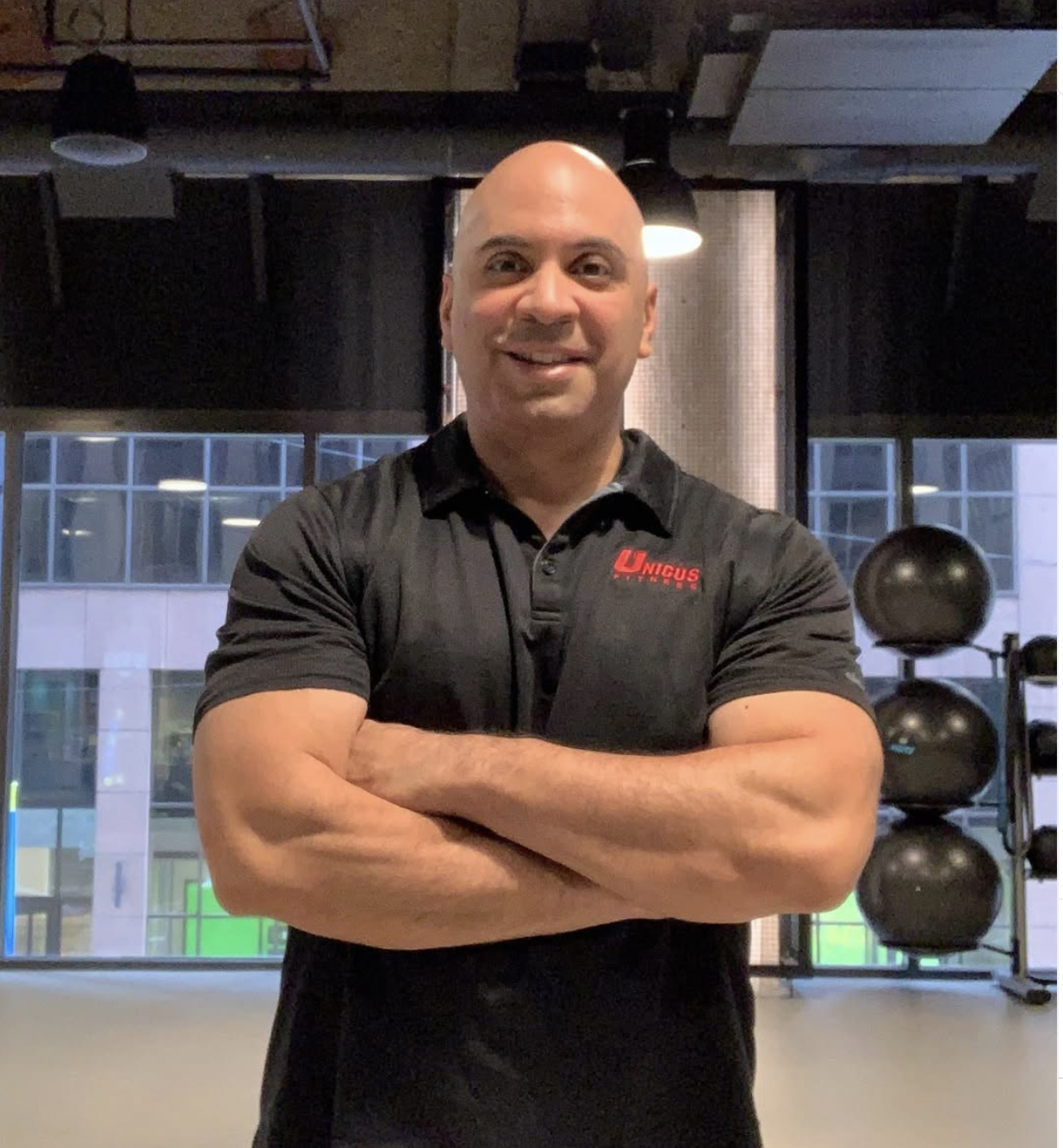 Frank Nunez, Elite Chicago Personal Trainer, smiling and standing with arms crossed, wearing a black polo shirt with red logo that reads 'Unicus Fitness', at his HQ in the Chicago Loop in the group fitness room.