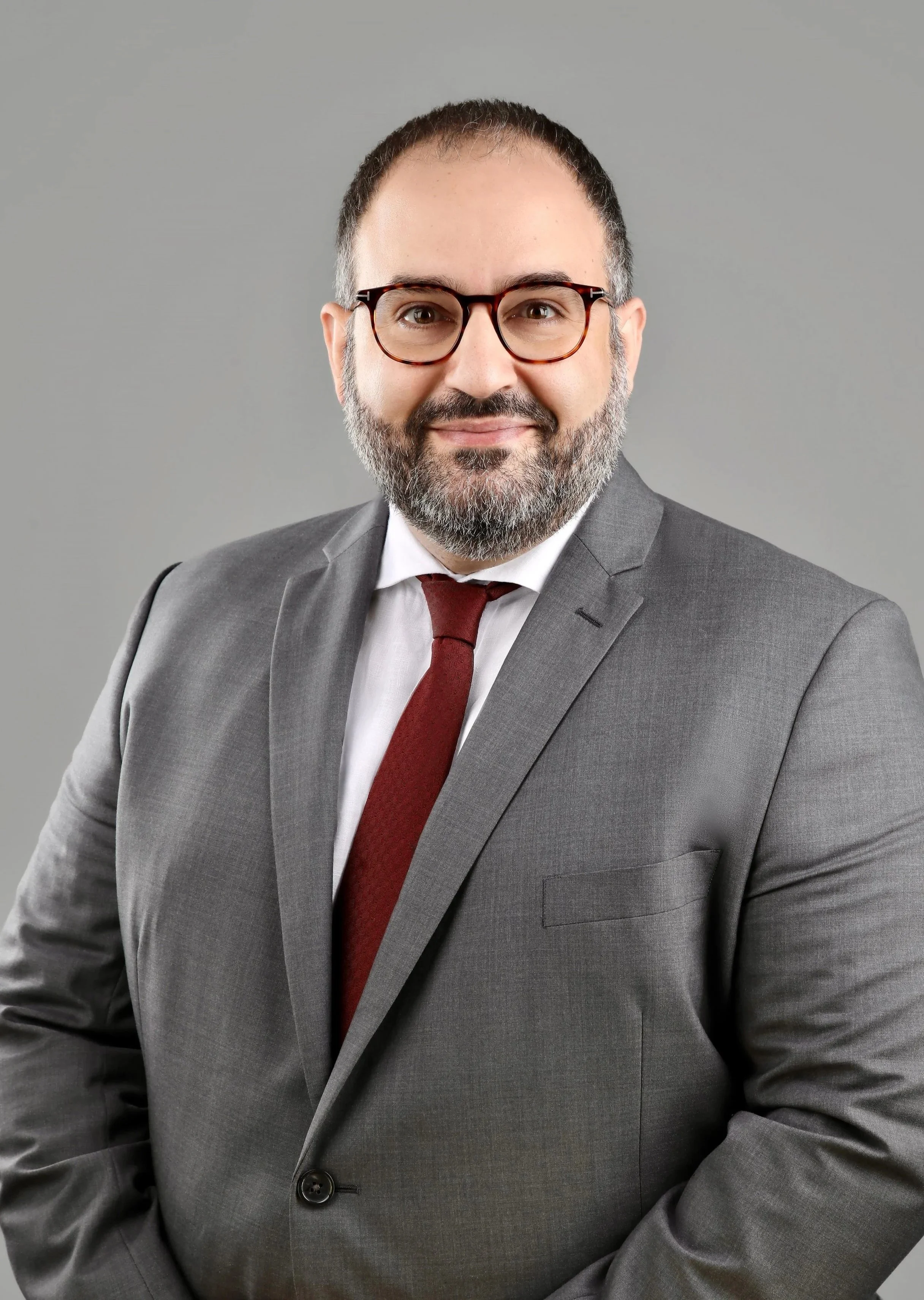 Headshot of a man in a gray suit, white shirt, maroon tie, with glasses, short dark hair, and a beard, smiling against a plain gray background.