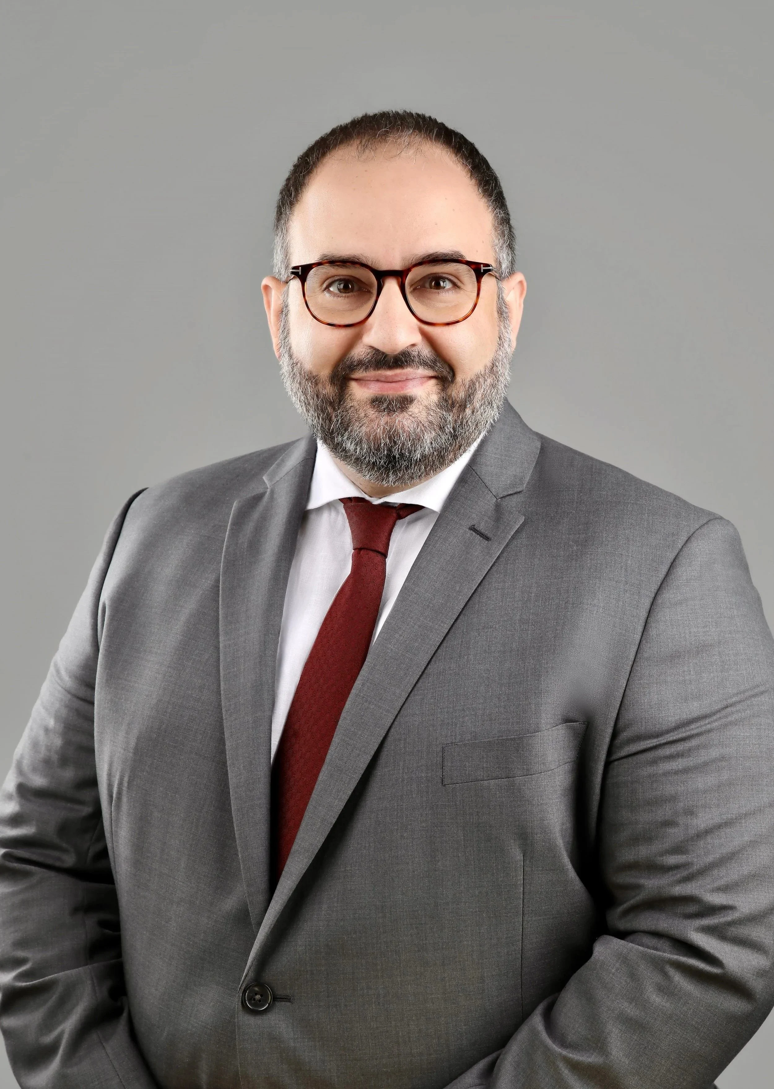 A man in a gray suit with a white shirt and red tie, wearing glasses, smiling against a gray background.