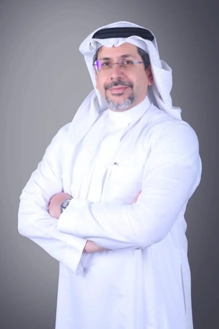 A man in traditional Middle Eastern attire, wearing a white thobe, a head covering, and glasses, standing with arms crossed and smiling.