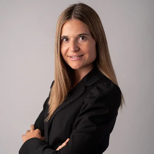 A woman with long brown hair, wearing a black blazer, smiling with arms crossed against a plain grey background.