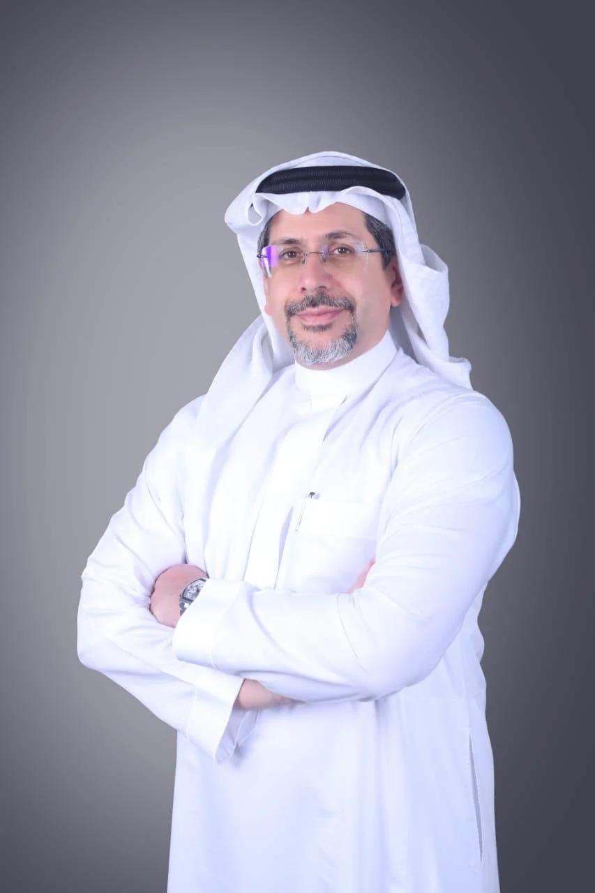 A man wearing traditional Middle Eastern attire, including a white thobe and head covering, standing with arms crossed against a gray background.