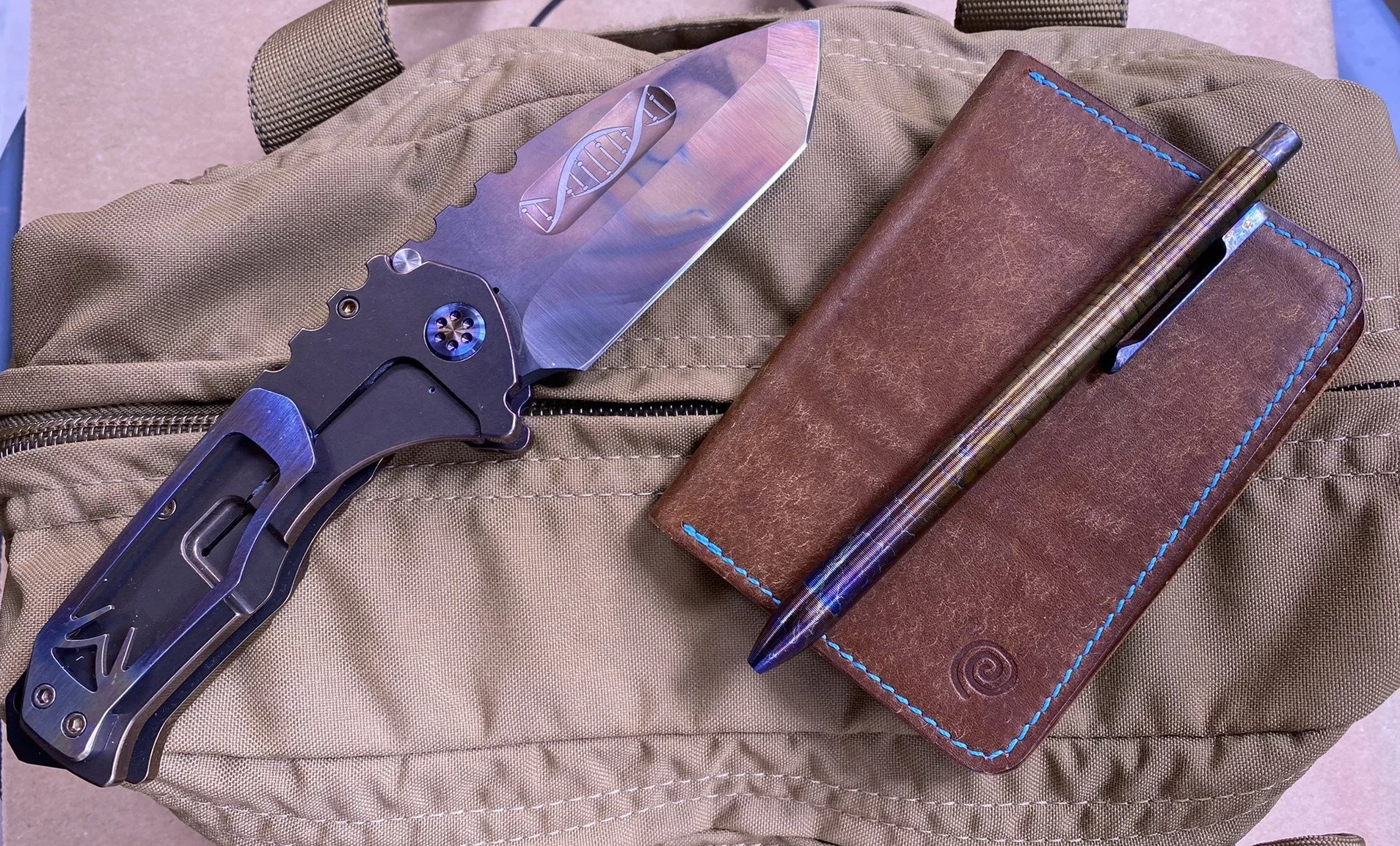 Medford Knife and Tool Praetorian TI knife, a Morgan Leatherworks leather-bound notebook, and a Tactile Turn metal pen resting on a tan fabric bag.