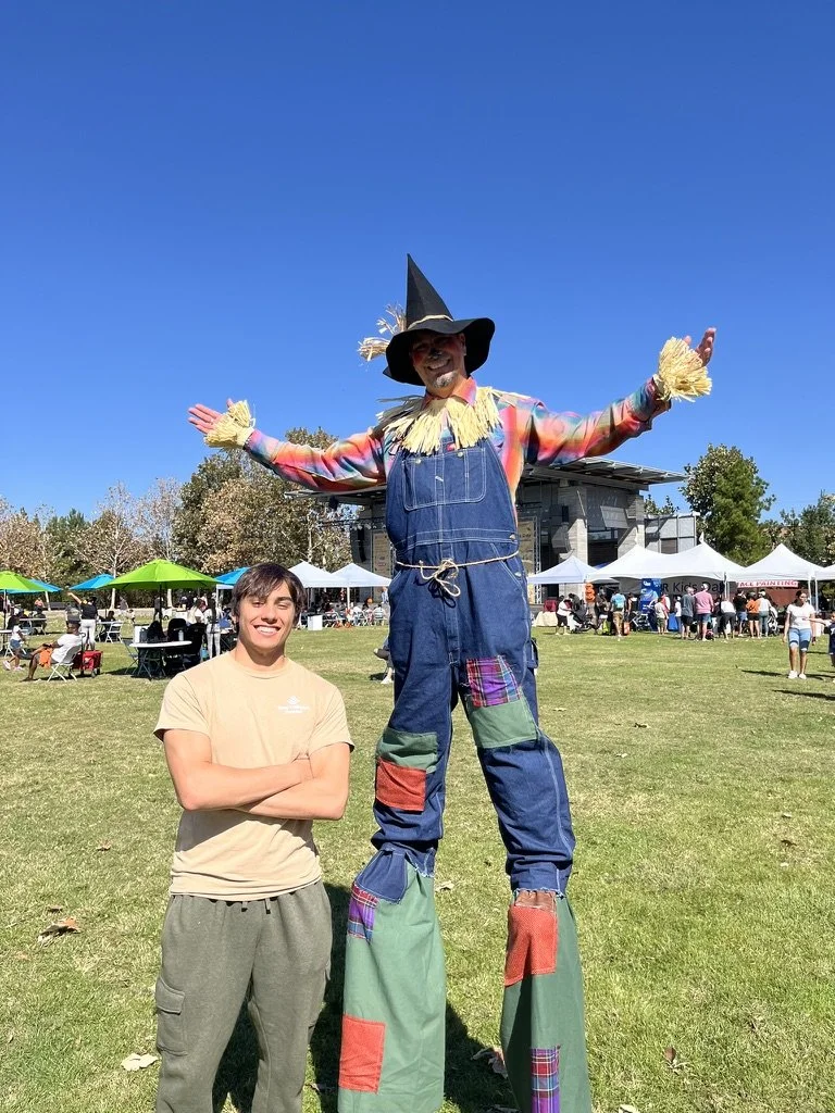 Man and a scarecrow at an outdoor event on a sunny day, with tents and people in the background.