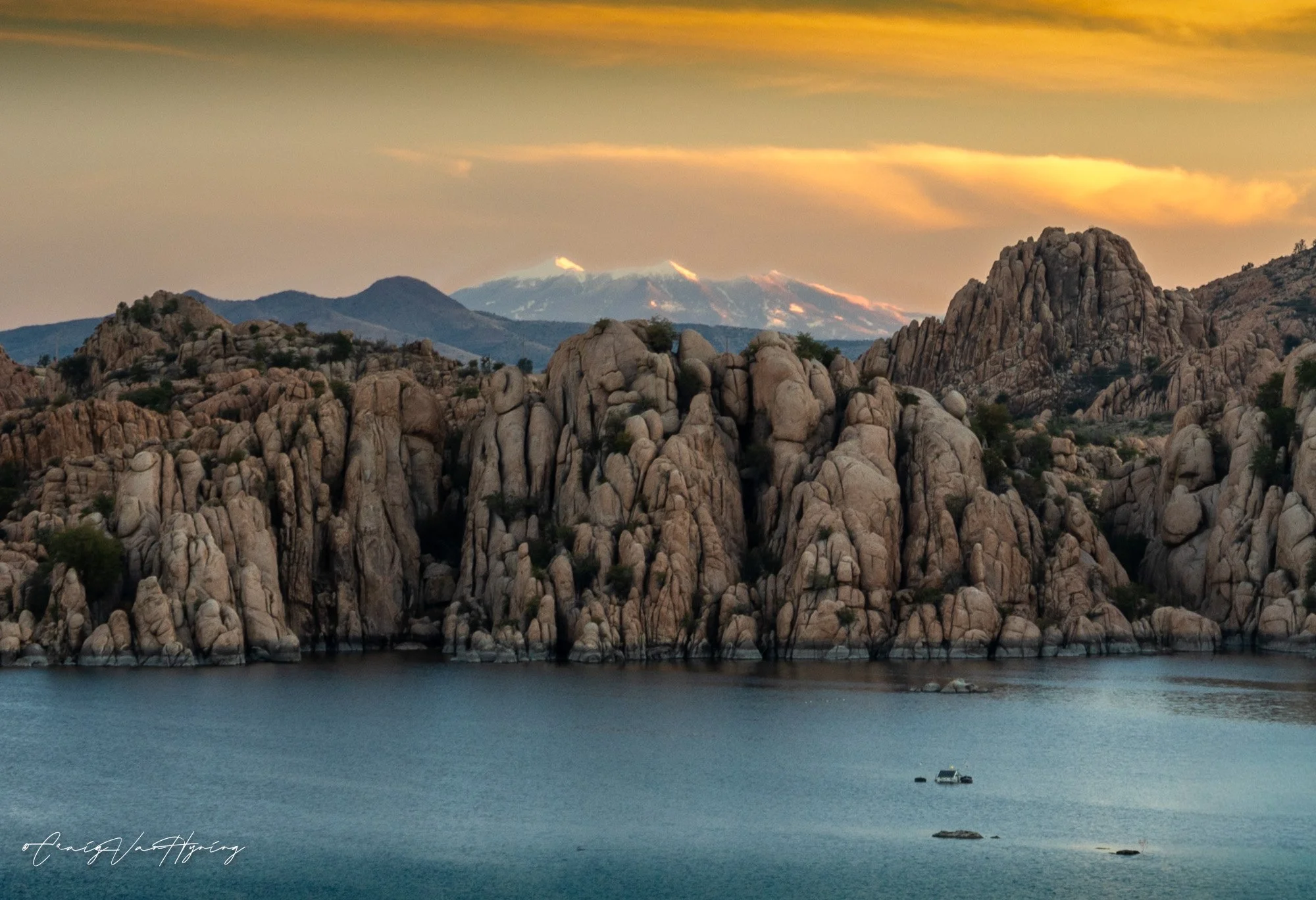 Scenic view of large rocky hills bordering a body of water at sunset, with mountains in the background and snow-capped peaks under a colored sky.