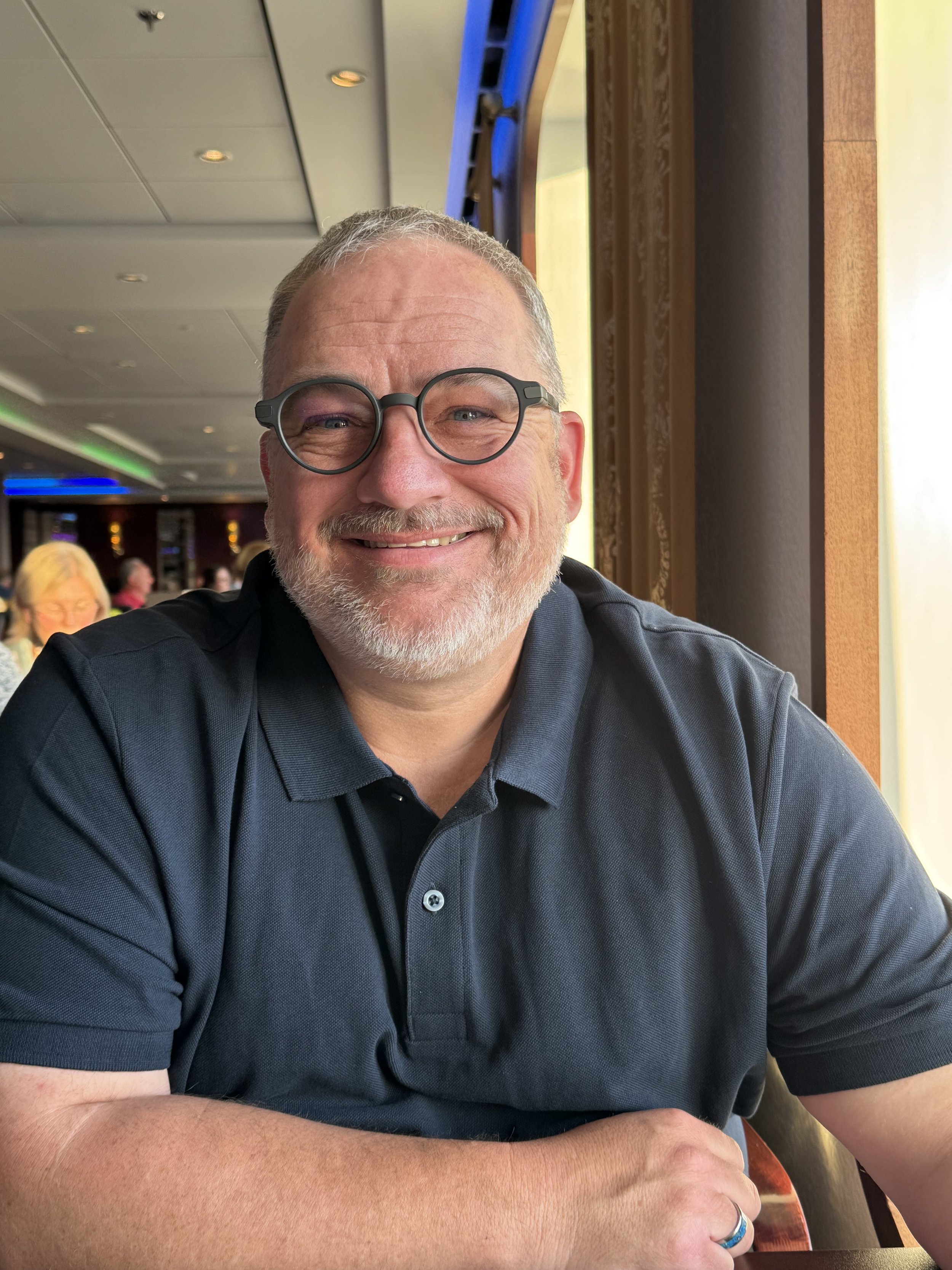 Craig himself with glasses, gray beard, smiling, sitting at a table by a window in a restaurant or cafe.