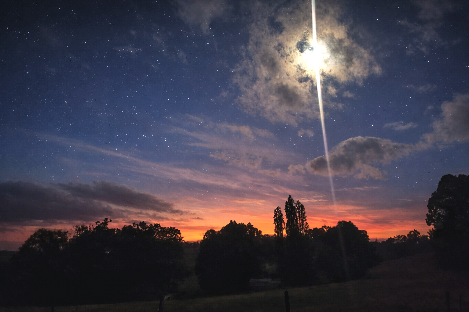 Night sky with stars and clouds, a moon partially covered by clouds. The horizon shows a sunset with orange and pink hues, and silhouettes of trees and fences in the foreground.