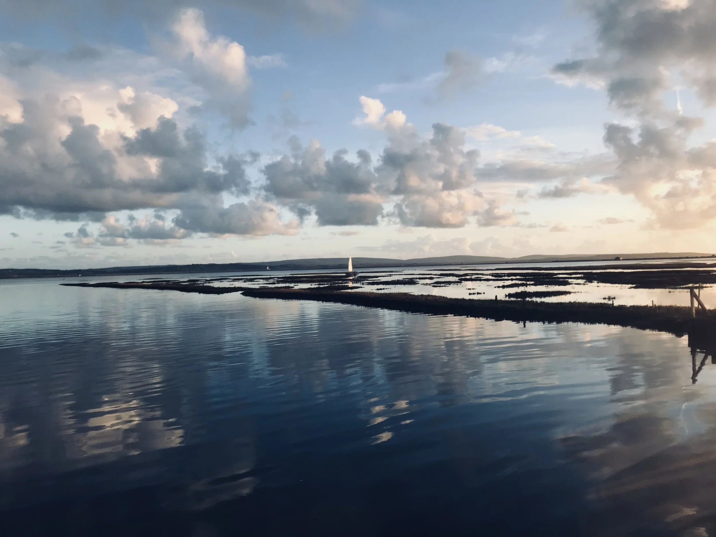 A serene body of water with reflections of clouds in the sky, featuring a sailboat and a distant shoreline during sunset.