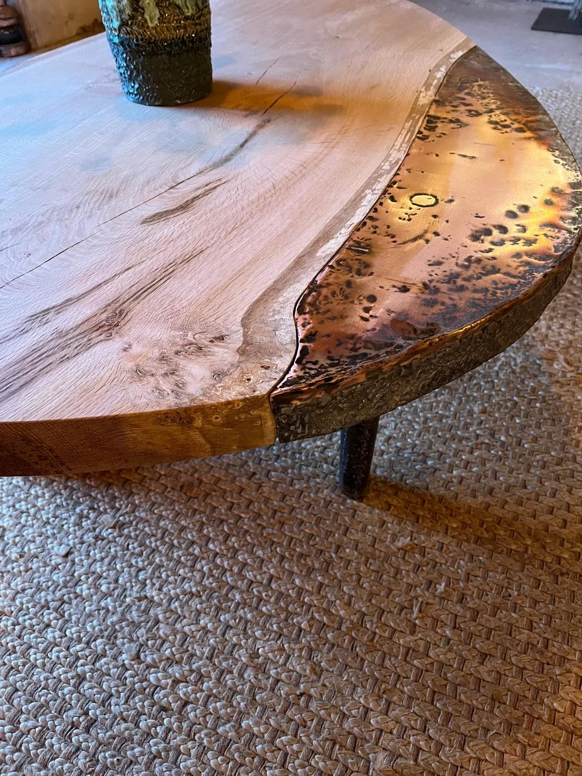 Close-up of a wooden table with a live edge, showing a mix of natural wood grain and a resin-filled border with a hammered metal finish. The table is placed on a woven carpet, and a glass with a textured surface is partially visible on the top left.