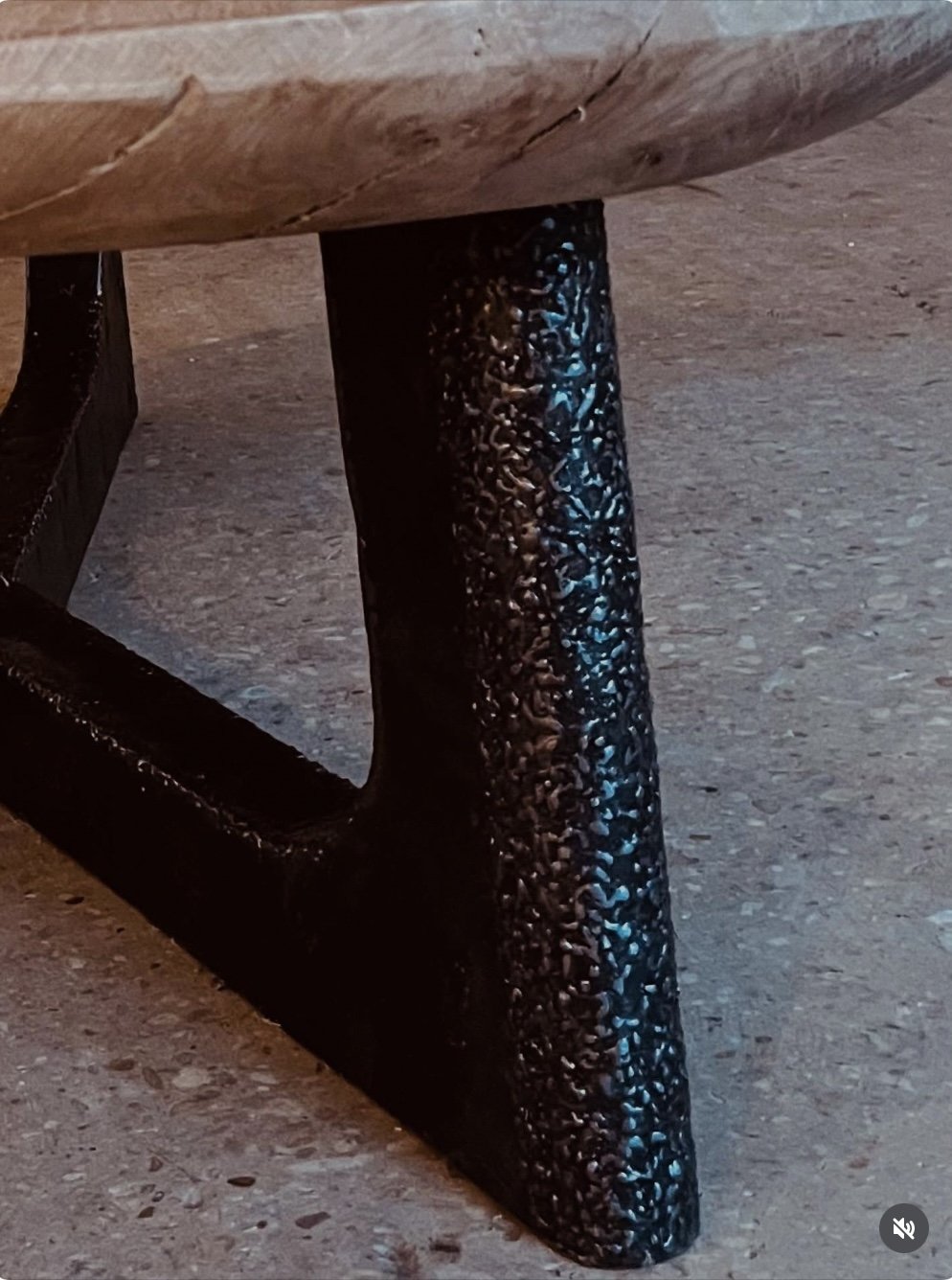 Close-up of a black, textured table leg supporting a wooden tabletop on a concrete floor.