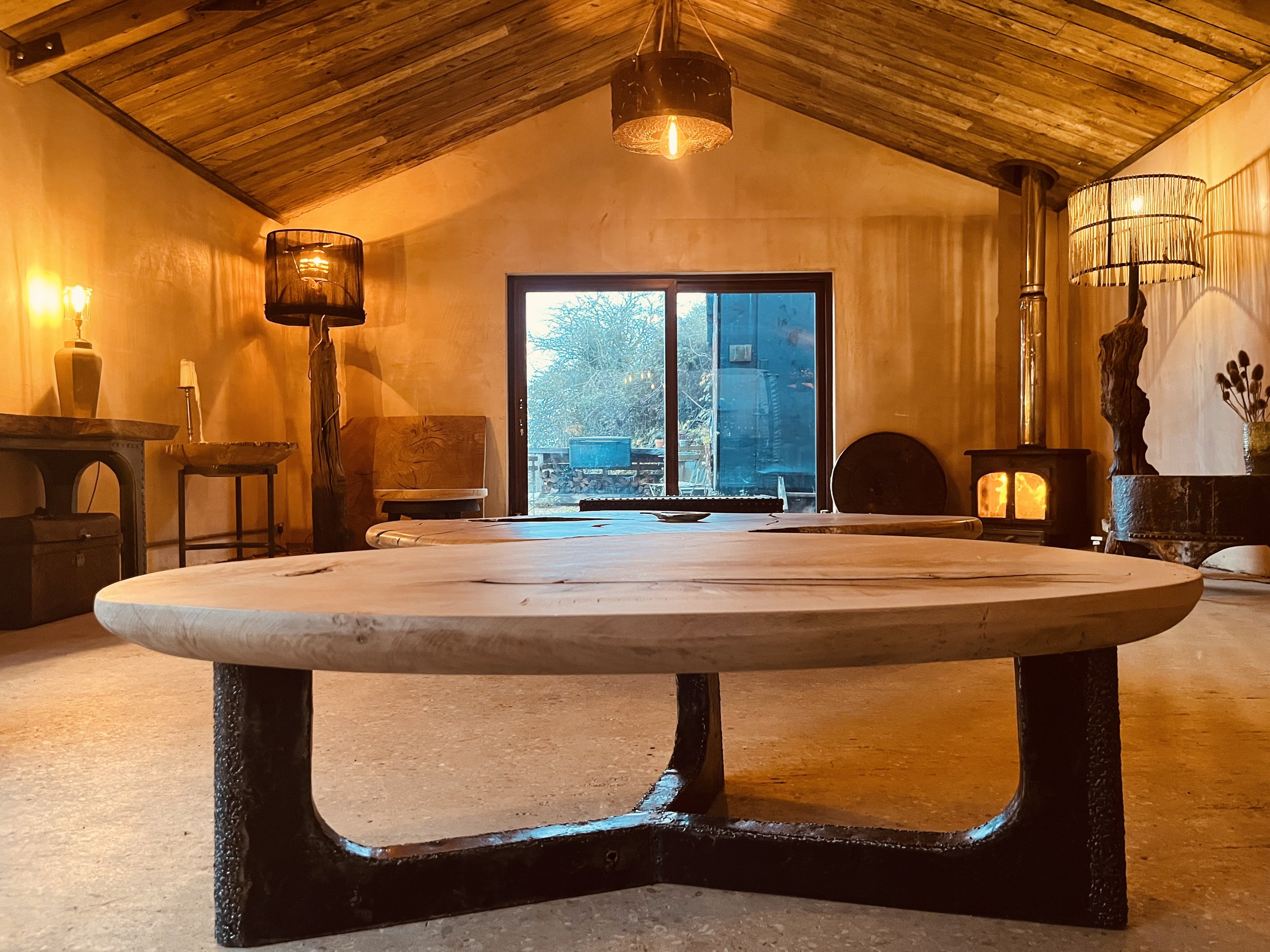 A cozy, warmly lit living room with wooden walls and ceiling, featuring a low, round wooden coffee table in the center, a wood stove emitting a soft glow, and various lamps providing ambient lighting, with a sliding glass door showing a view of trees outside.