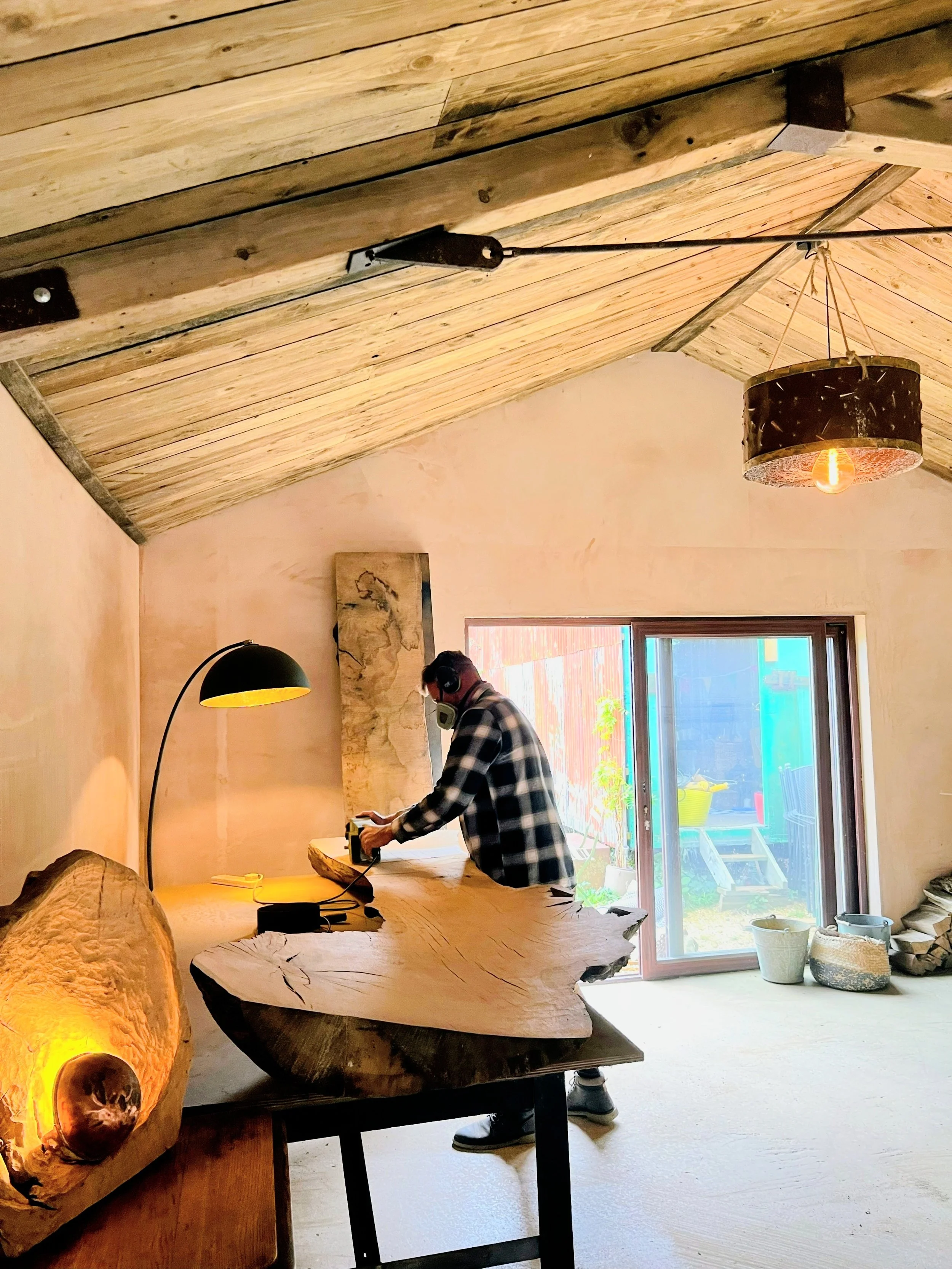 A person in a plaid shirt and safety mask working with a power tool on a large, natural wood slab in a woodworking or carpentry workshop. The room has a sloped wooden ceiling, a sliding glass door letting in natural light, and decorative lamps.