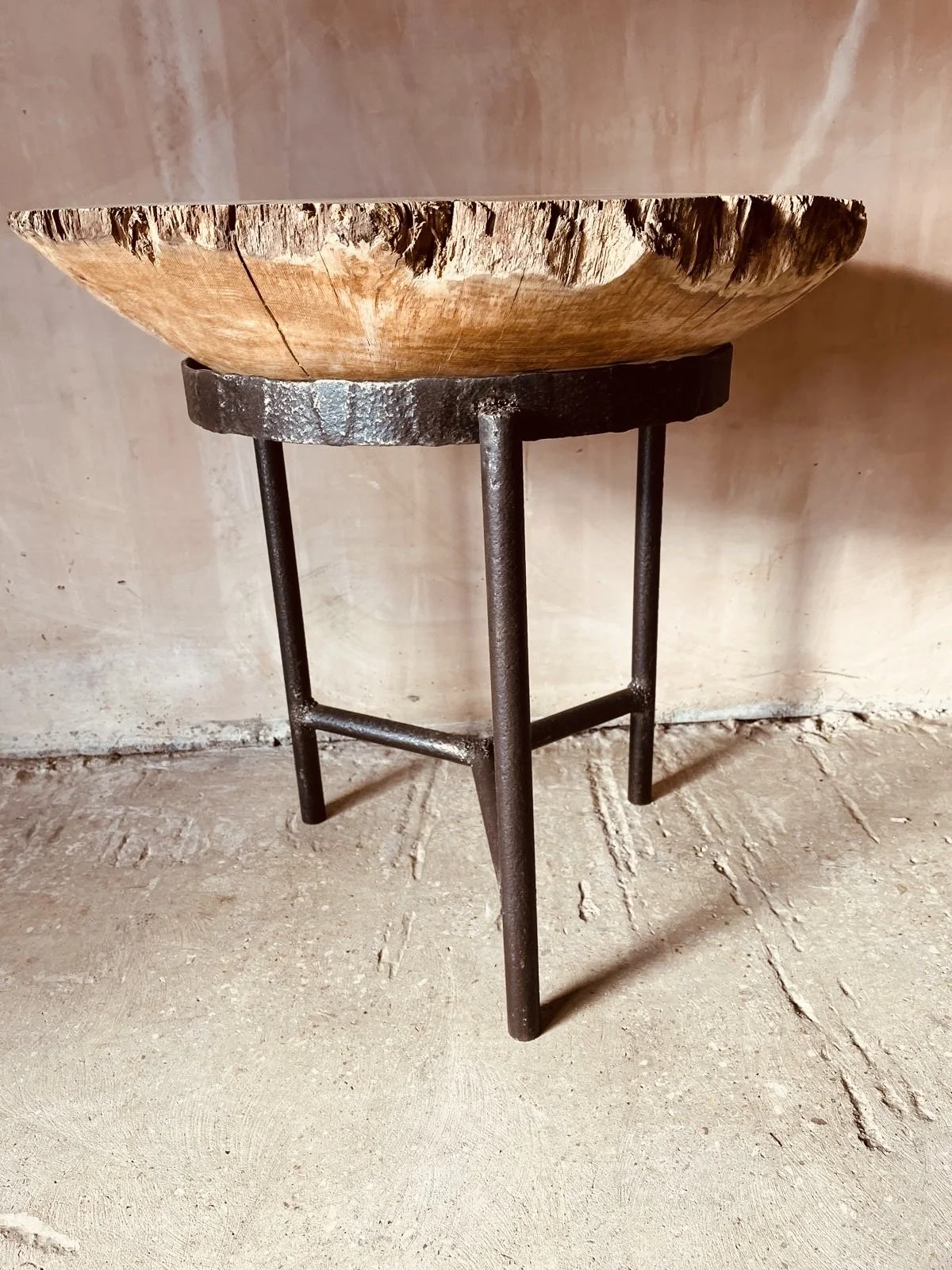 A wooden bowl on a metal stand with three legs, placed on a concrete floor against a plain wall.