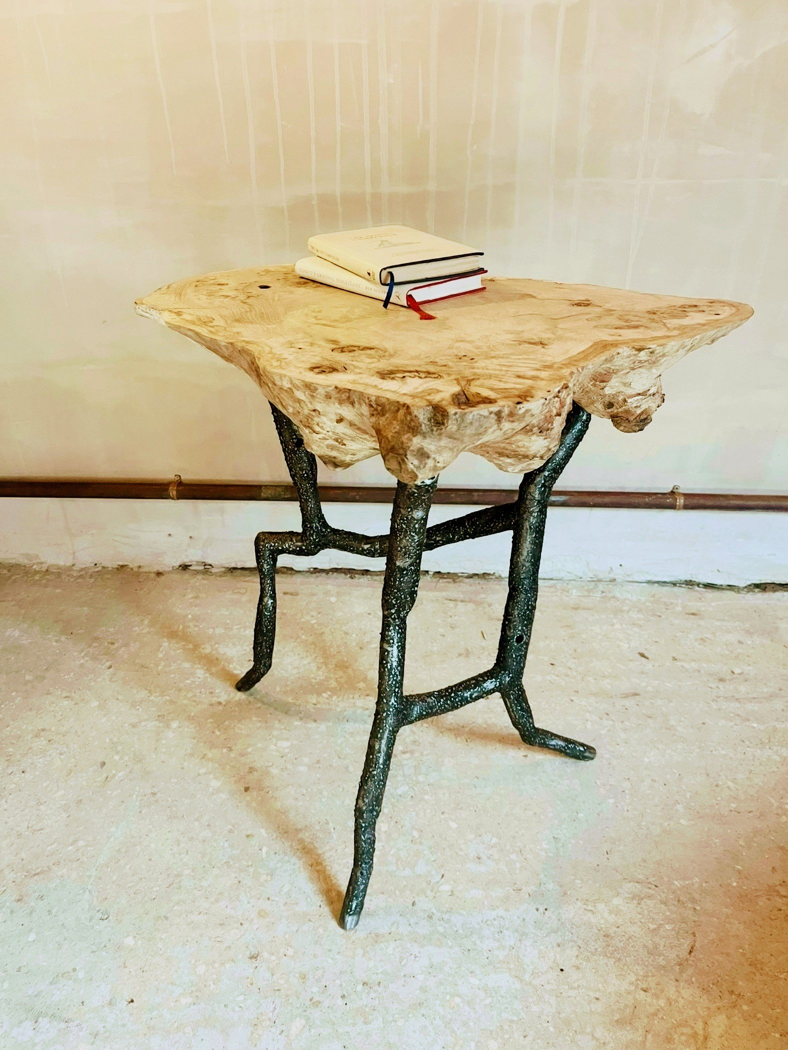 A rustic side table with a natural wood slab top and metal branch-like legs, with three books placed on top.