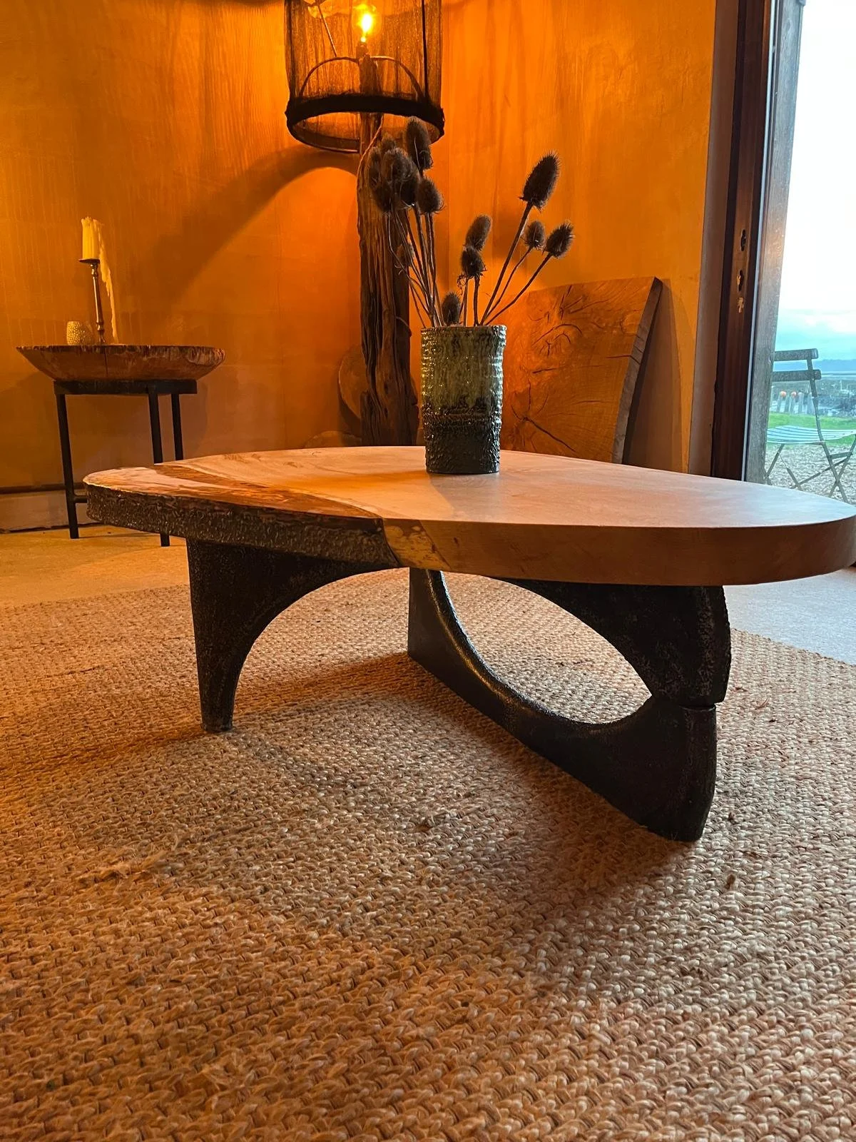 A rustic wooden coffee table with a curved black base, placed on a woven rug, with a green vase containing dried flowers on top. In the background, there's a wooden wall and a side table with a candle, along with a decorative lamp and a piece of wooden art near a glass door leading outside.