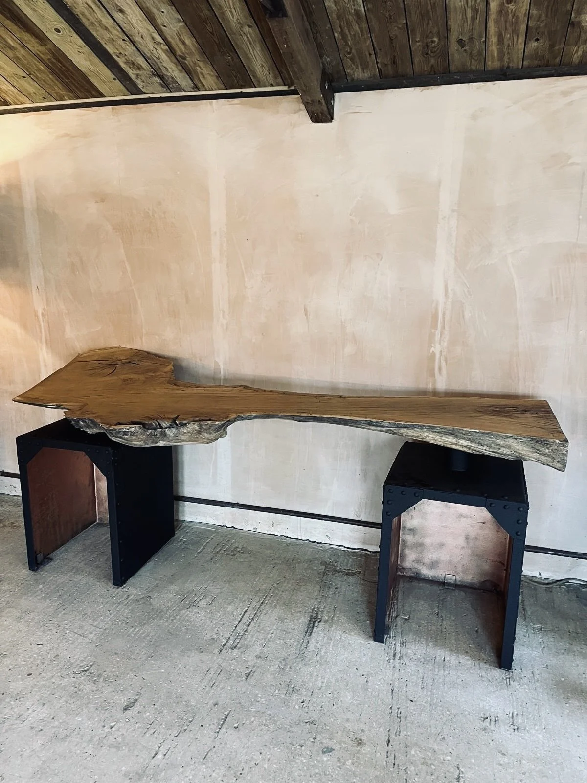 A rustic wooden table with a live edge design, supported by two black modern geometric stands, set against a minimalist wall and ceiling with wood paneling.