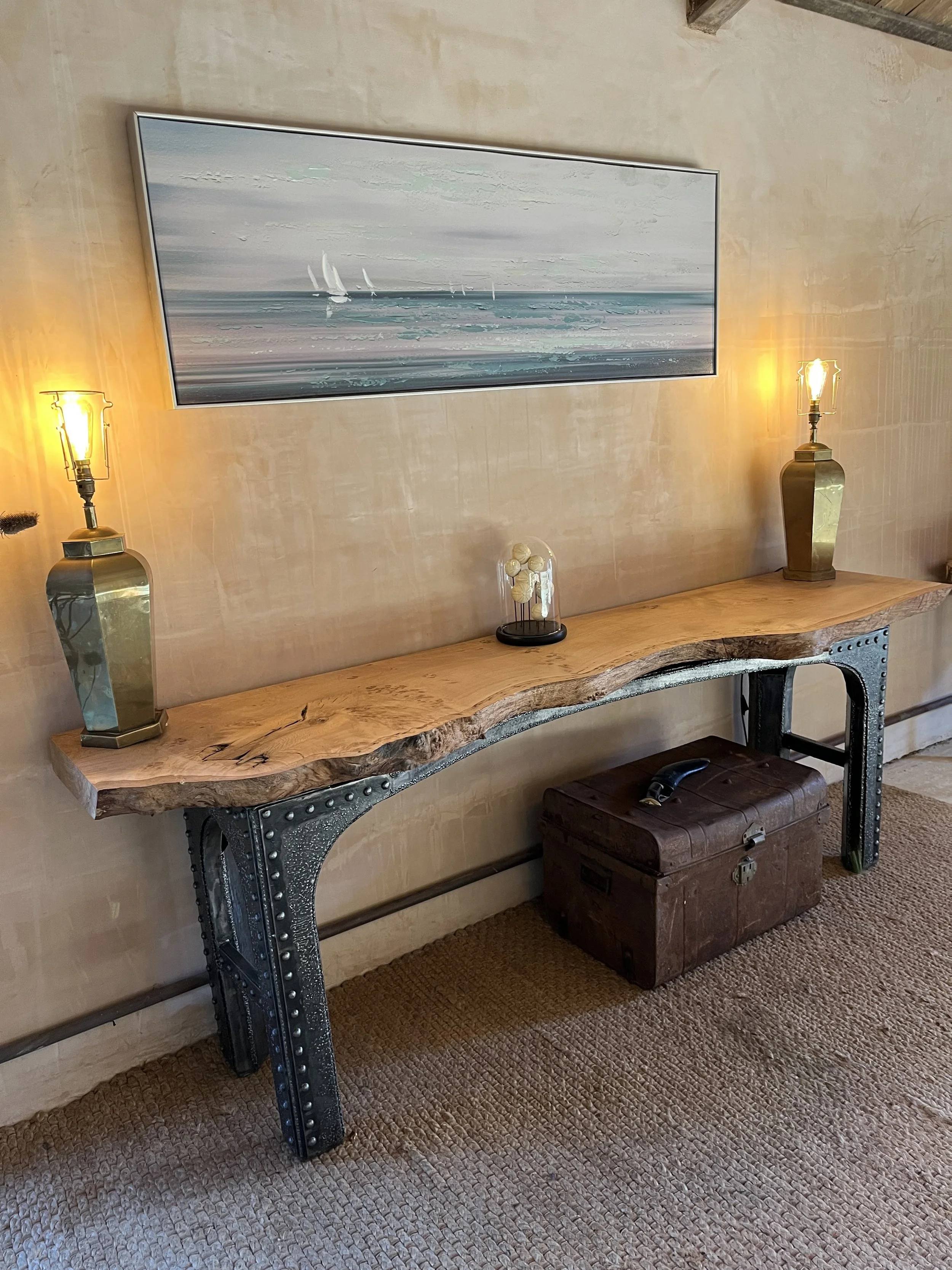 A wall-mounted wooden console table with metal legs, two gold lamps, a glass dome with decorative white objects, and a vintage trunk underneath. A large landscape painting of a seascape with sailboats hangs above.