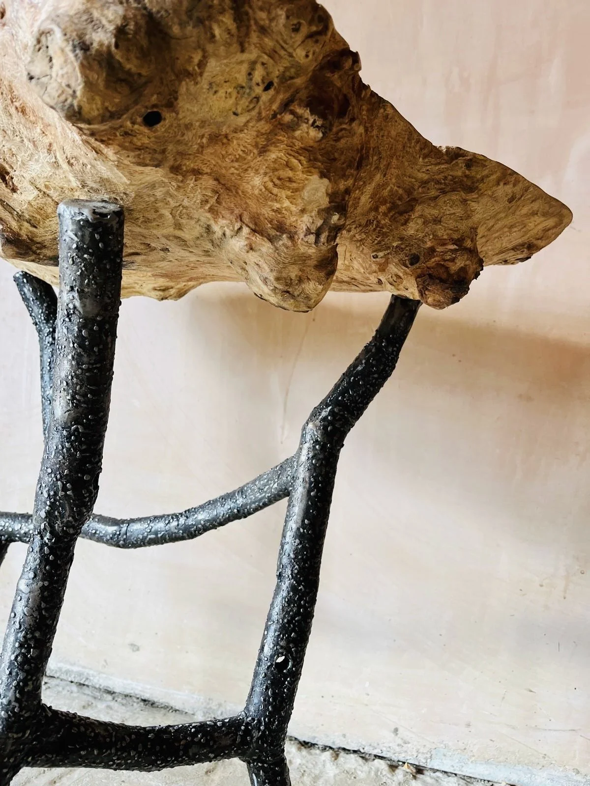 Close-up of a wooden chair with a seat made from a natural wood slab and black metal legs with textured droplets on them, set against a light-colored wall.