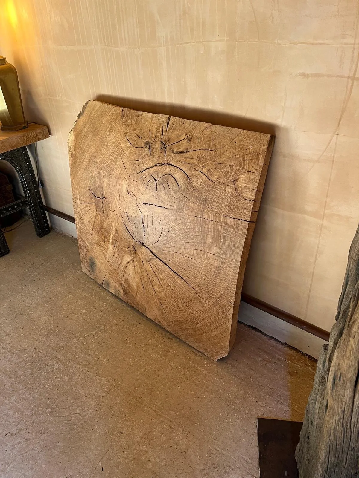 A large square wooden slab with visible tree rings and cracks leaning against a beige wall. Part of a black and wooden furniture piece and a small wooden side table are also visible.