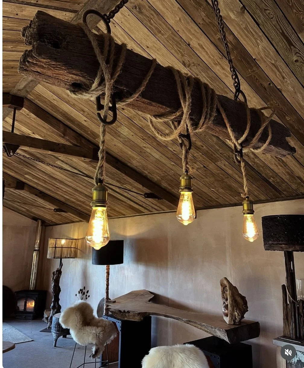 Rustic interior with wooden ceiling, hanging rope and log light fixtures, Edison bulbs, and modern furniture including furry chairs and lamps.