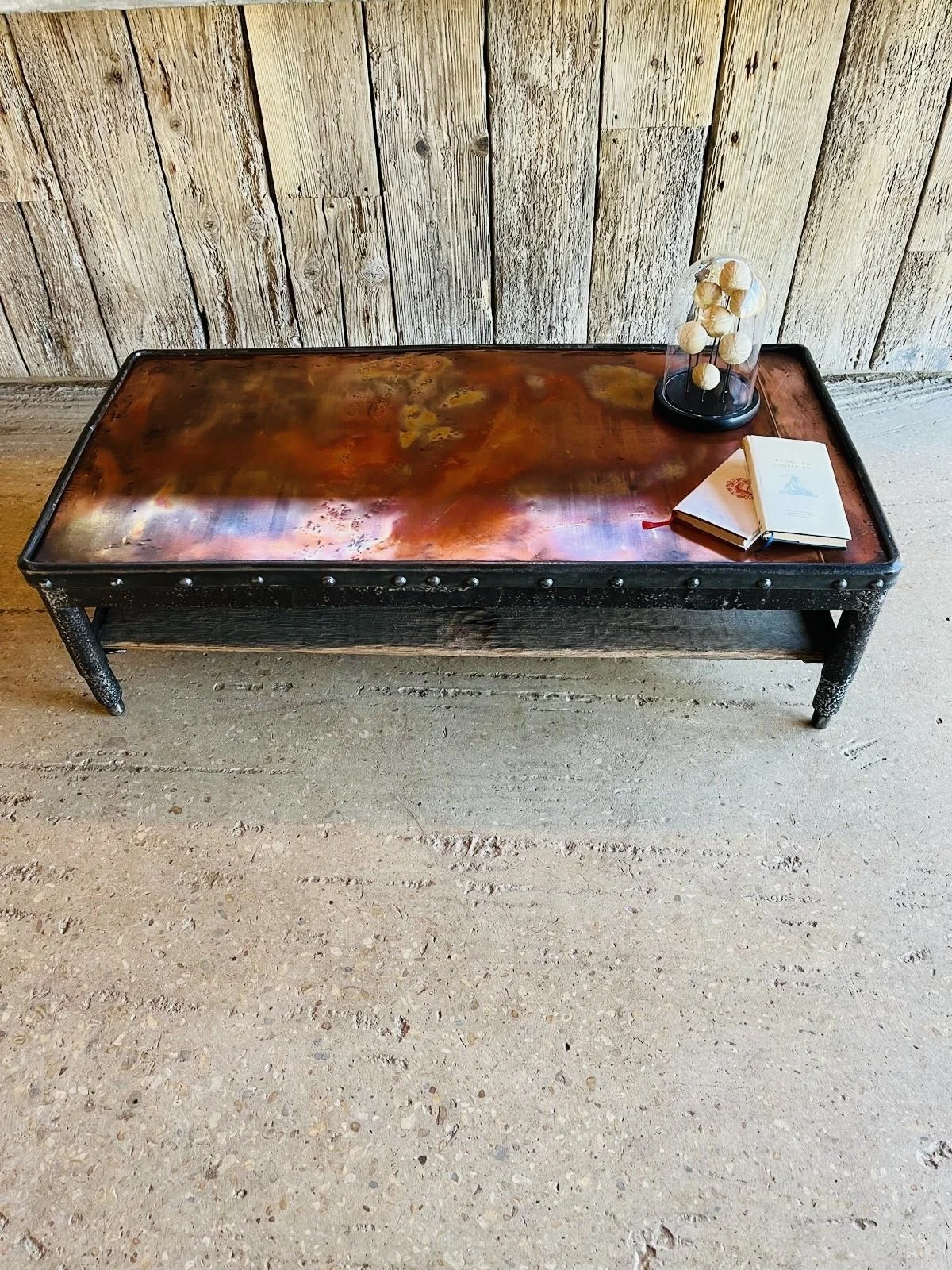 A rustic wooden and metal coffee table with a distressed surface, placed on a concrete floor against a weathered wooden wall. The table has decorative nailhead trim along the edges, and on it are a small black display dome containing white spherical objects, a closed book, and an open notebook with a red ribbon bookmark.